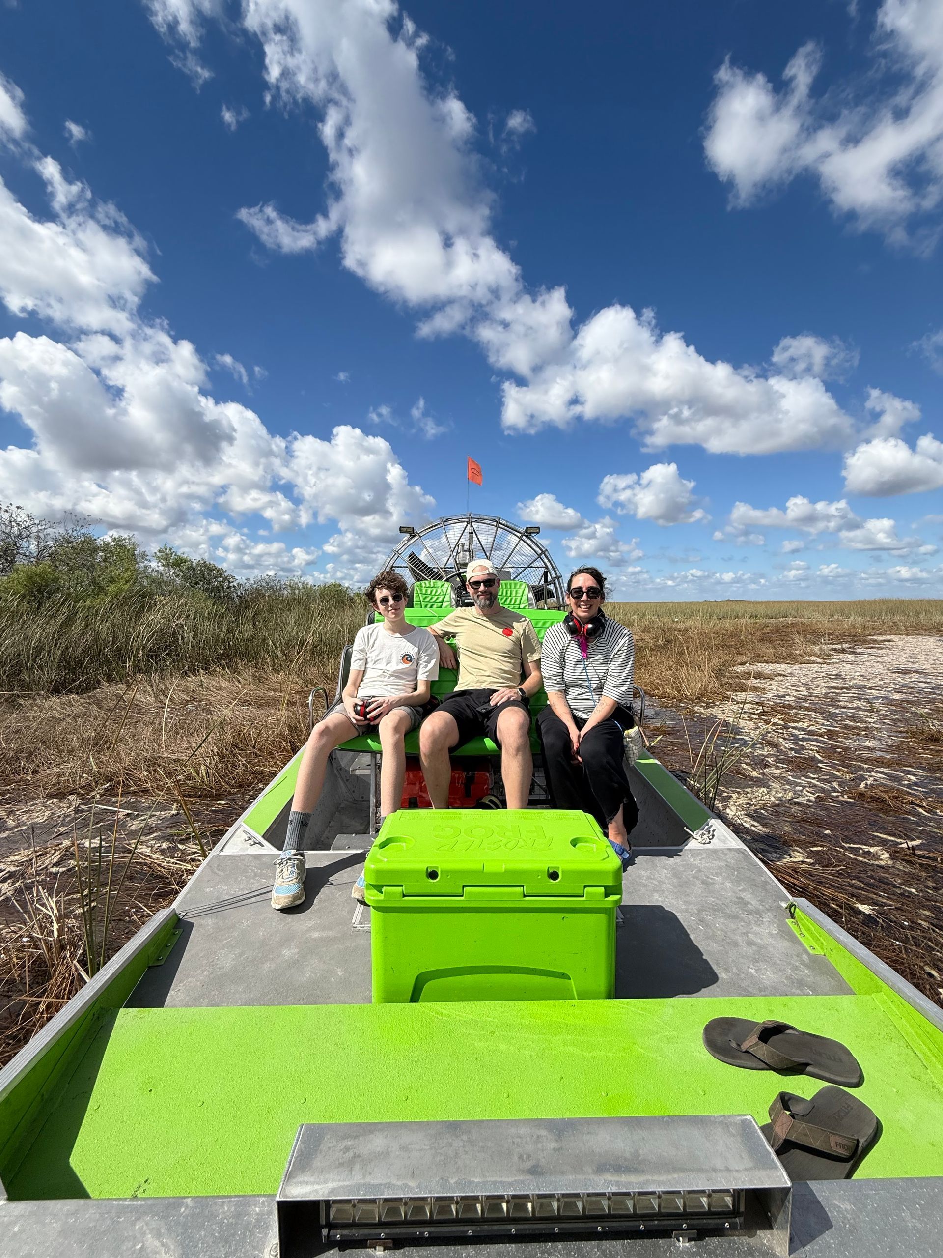 Three people on a green airboat in the Everglades under a blue sky with clouds.