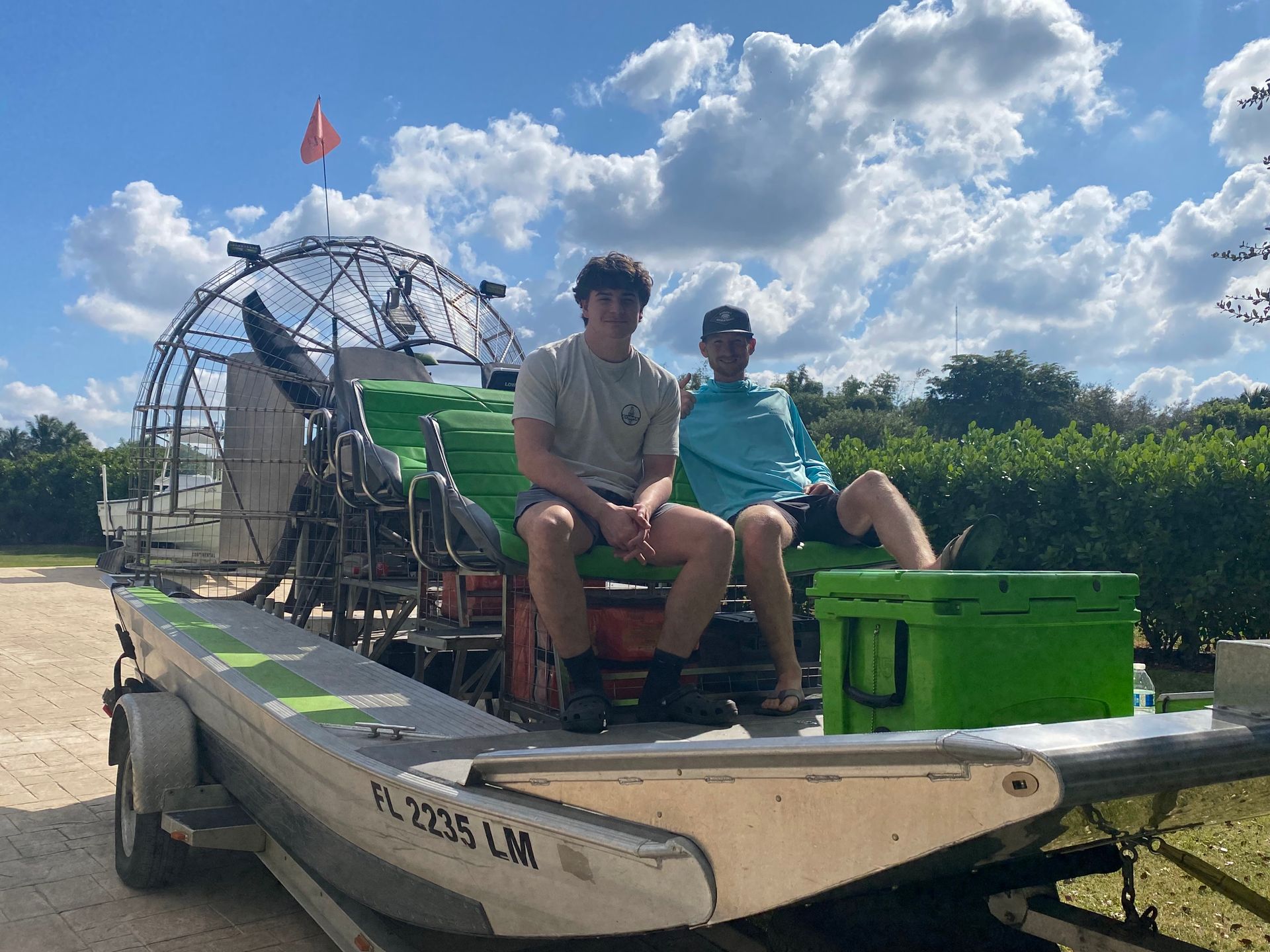 Two people sit on an airboat trailer, under a blue sky. The airboat is green and silver.