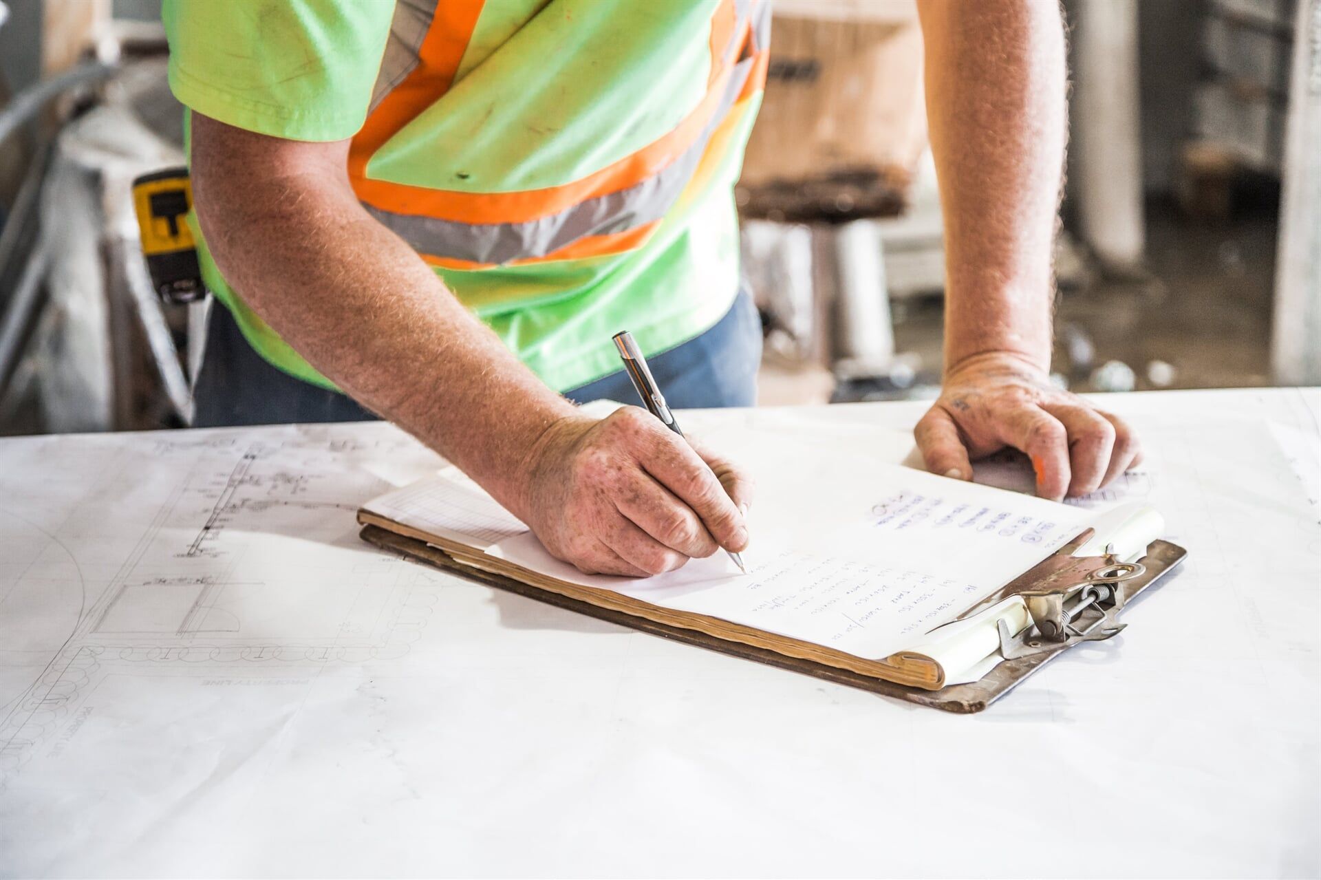 Construction worker in a green vest, writing on a clipboard resting on a large blueprint. — Kareela Constructions In Tamworth, NSW