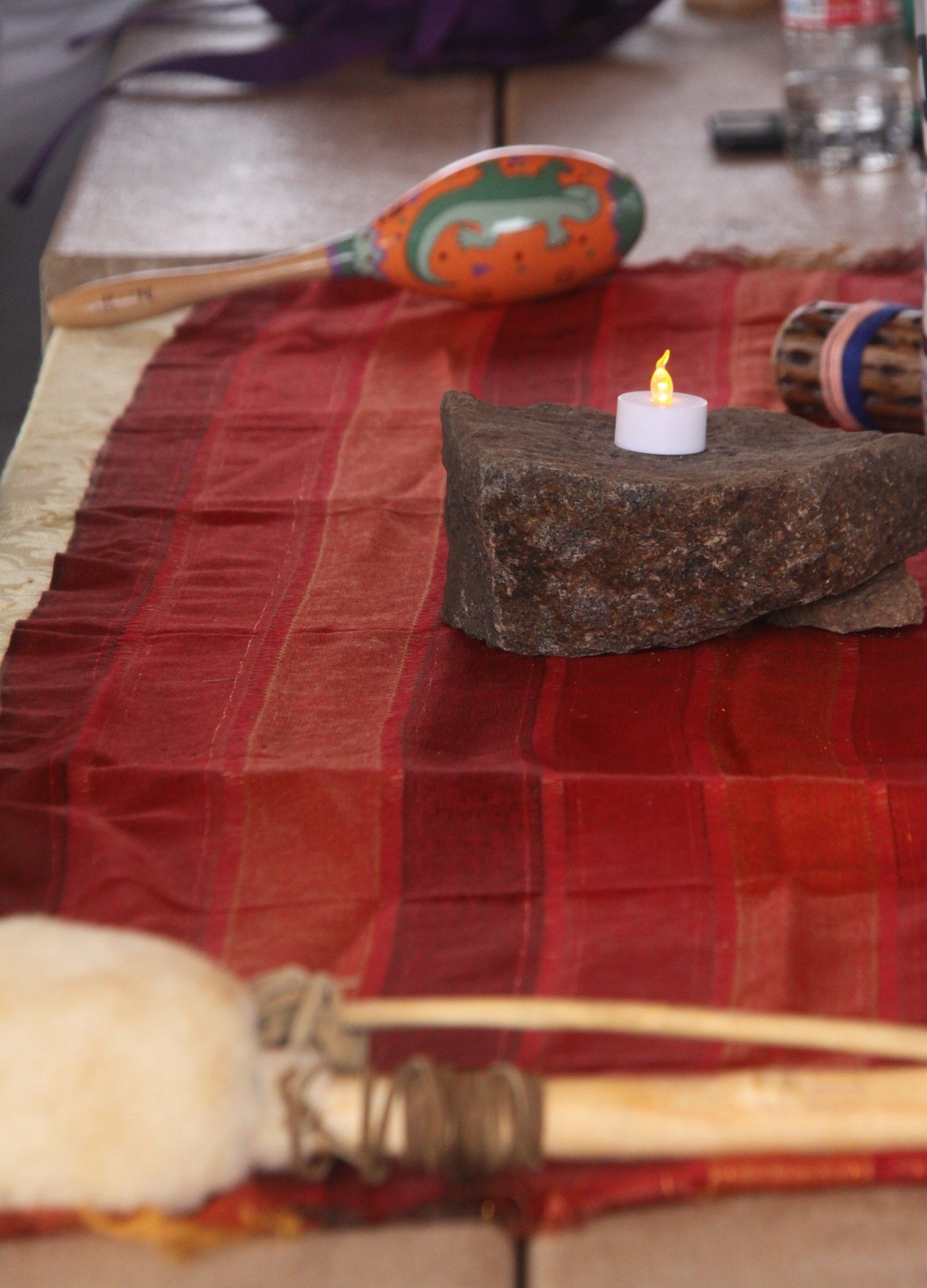 Red woven cloth with a candle on a stone, maracas, and a drum mallet on a table.
