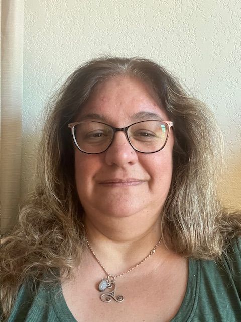 Woman with glasses smiling, wearing a green shirt and necklace, in front of a light-colored wall.