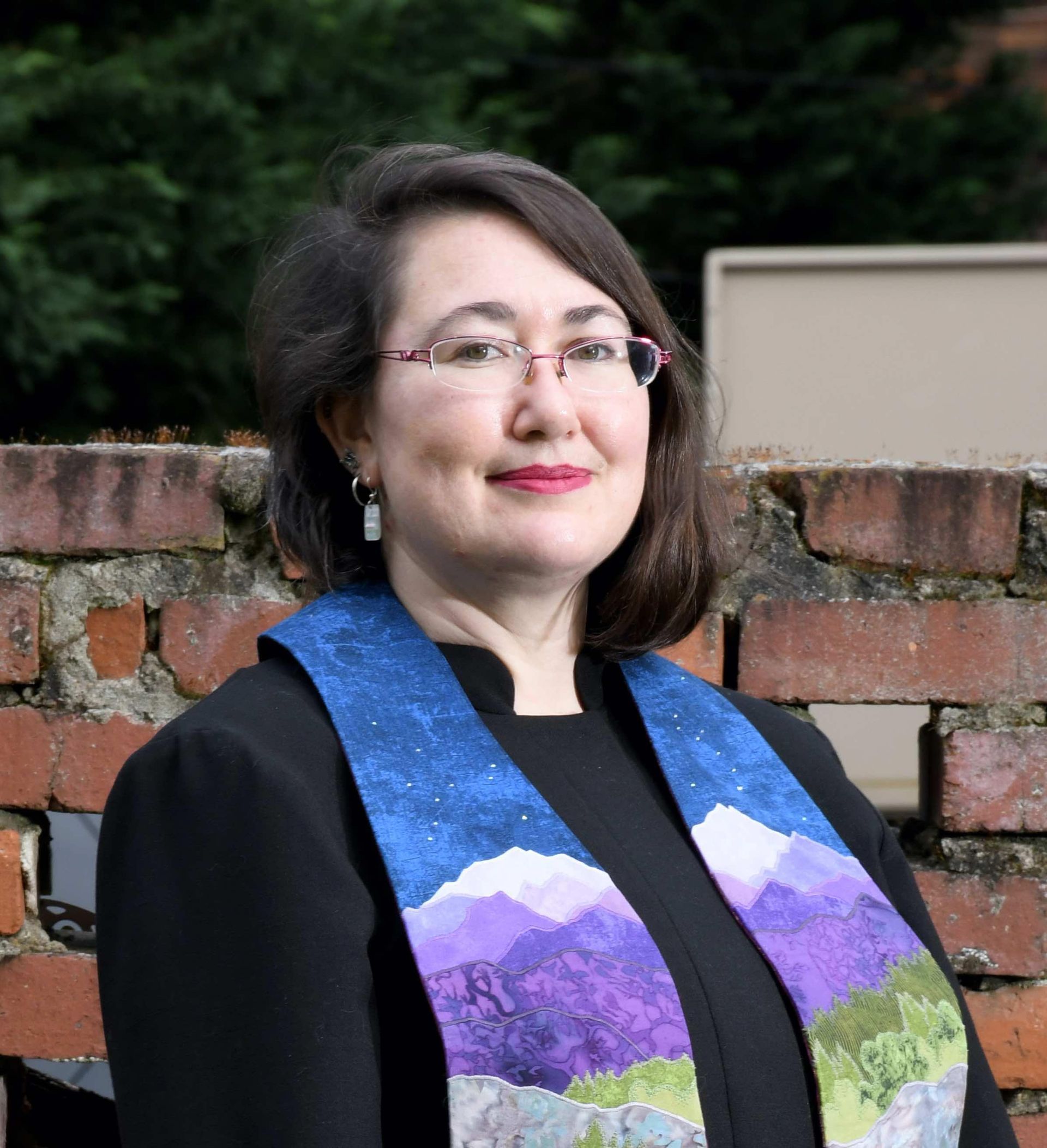 Woman with glasses and a colorful stole stands in front of a brick wall, smiling.