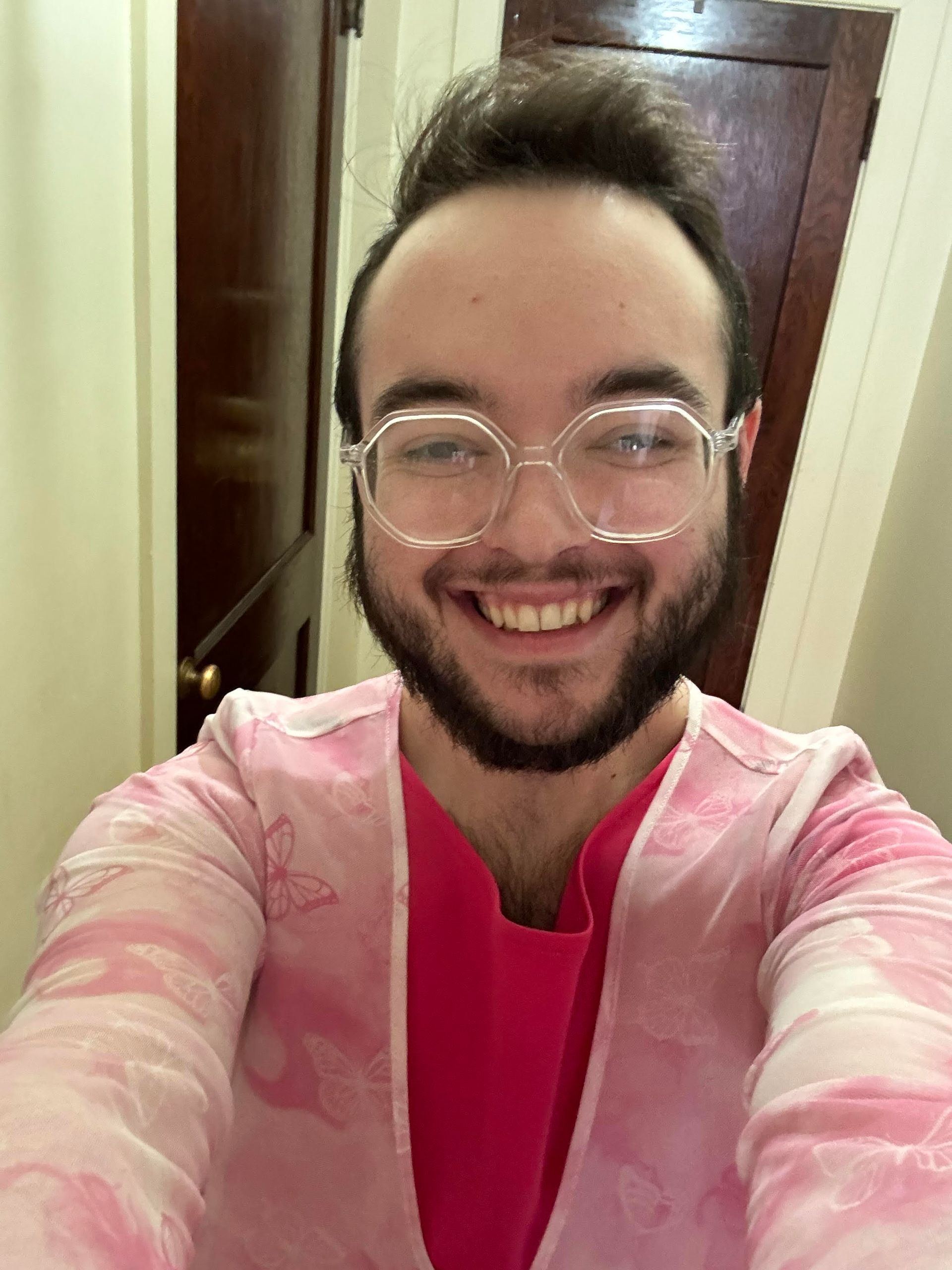 A smiling person wearing glasses and a pink outfit stands in a hallway, looking at the camera.