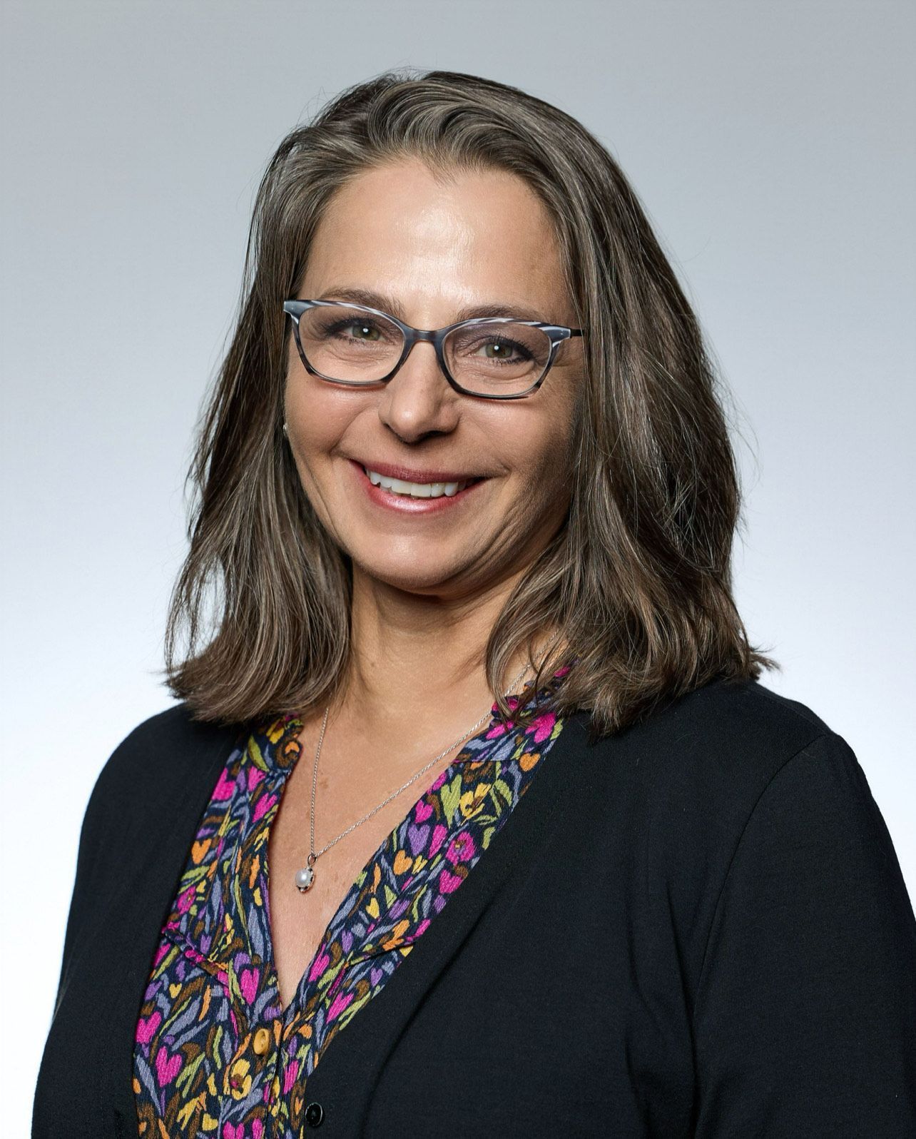 Woman with glasses smiles, wearing a patterned top under a black cardigan, against a grey background.