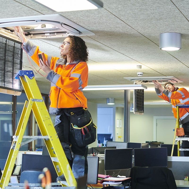 Commercial Electrical Contractor - A woman is standing on a yellow ladder working on an air conditioner