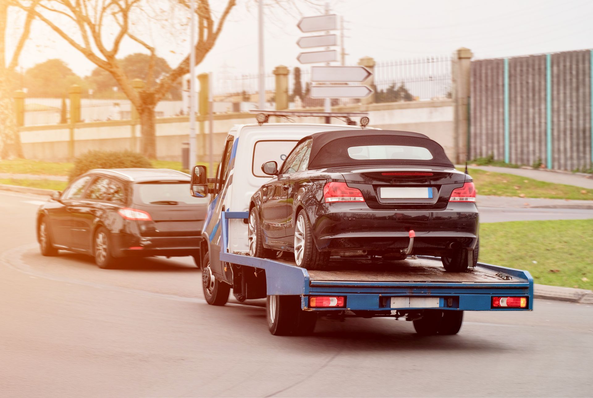 Tow truck carrying a black convertible on a flatbed, a car behind it on a sunny road.