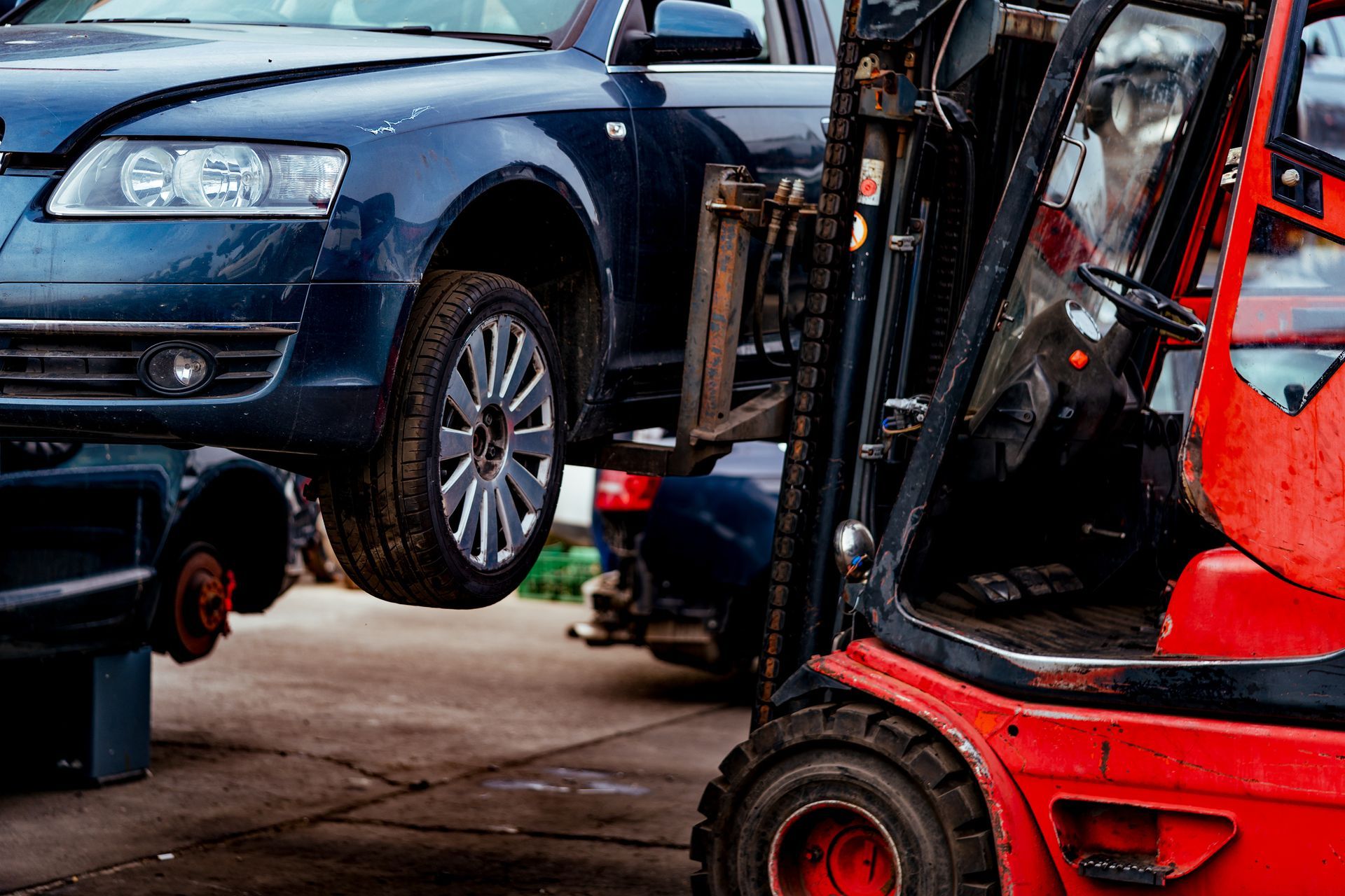 Red forklift lifting a blue car in a junkyard; wheel is off the car.