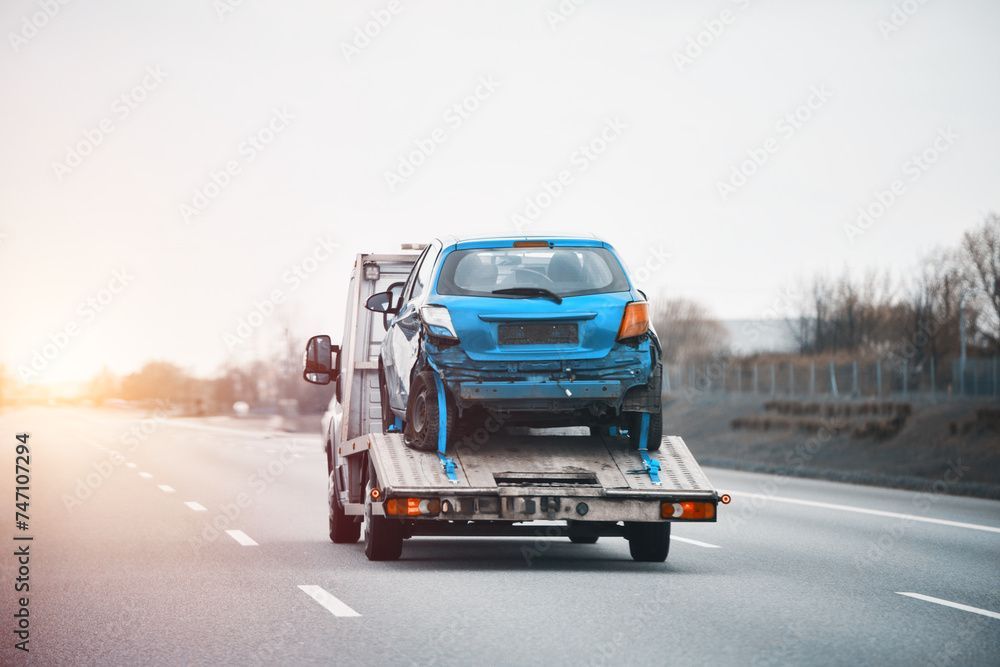Blue damaged car on a tow truck, traveling on a highway.