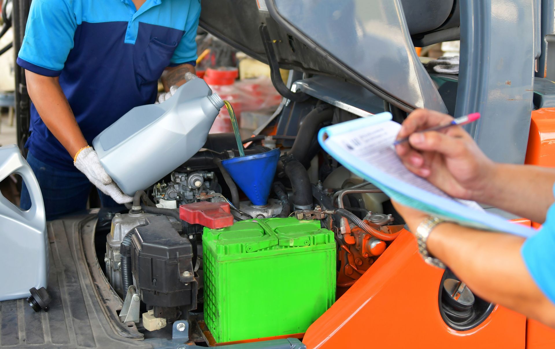 Mechanic pours oil into a forklift engine, while another checks a clipboard.