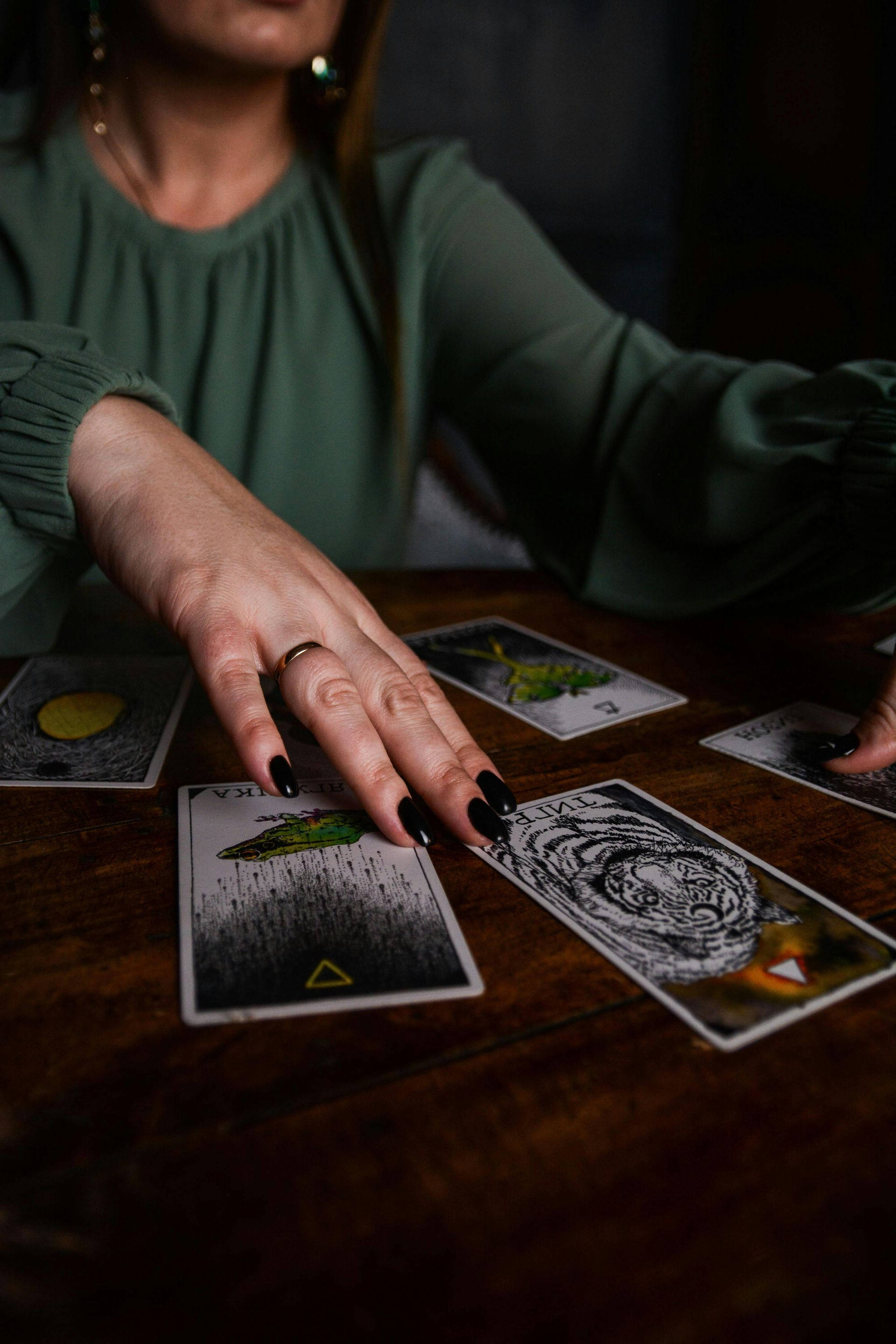 Person's hand over tarot cards on a wooden table, wearing a green blouse and black nail polish.