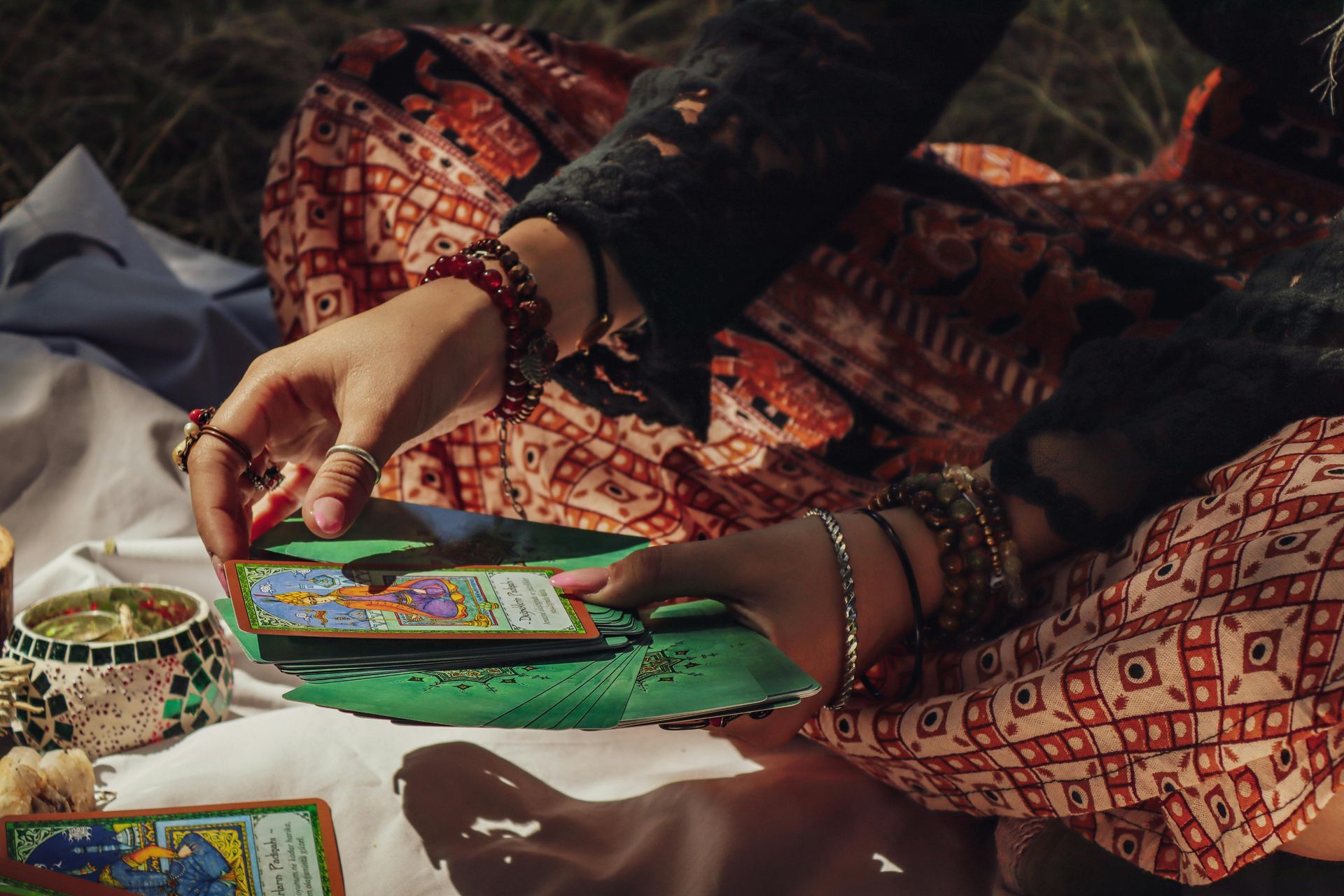 Person holding tarot cards, outdoors. Cards are laid on a green mat on white cloth. Colorful clothing, candles, bracelets.