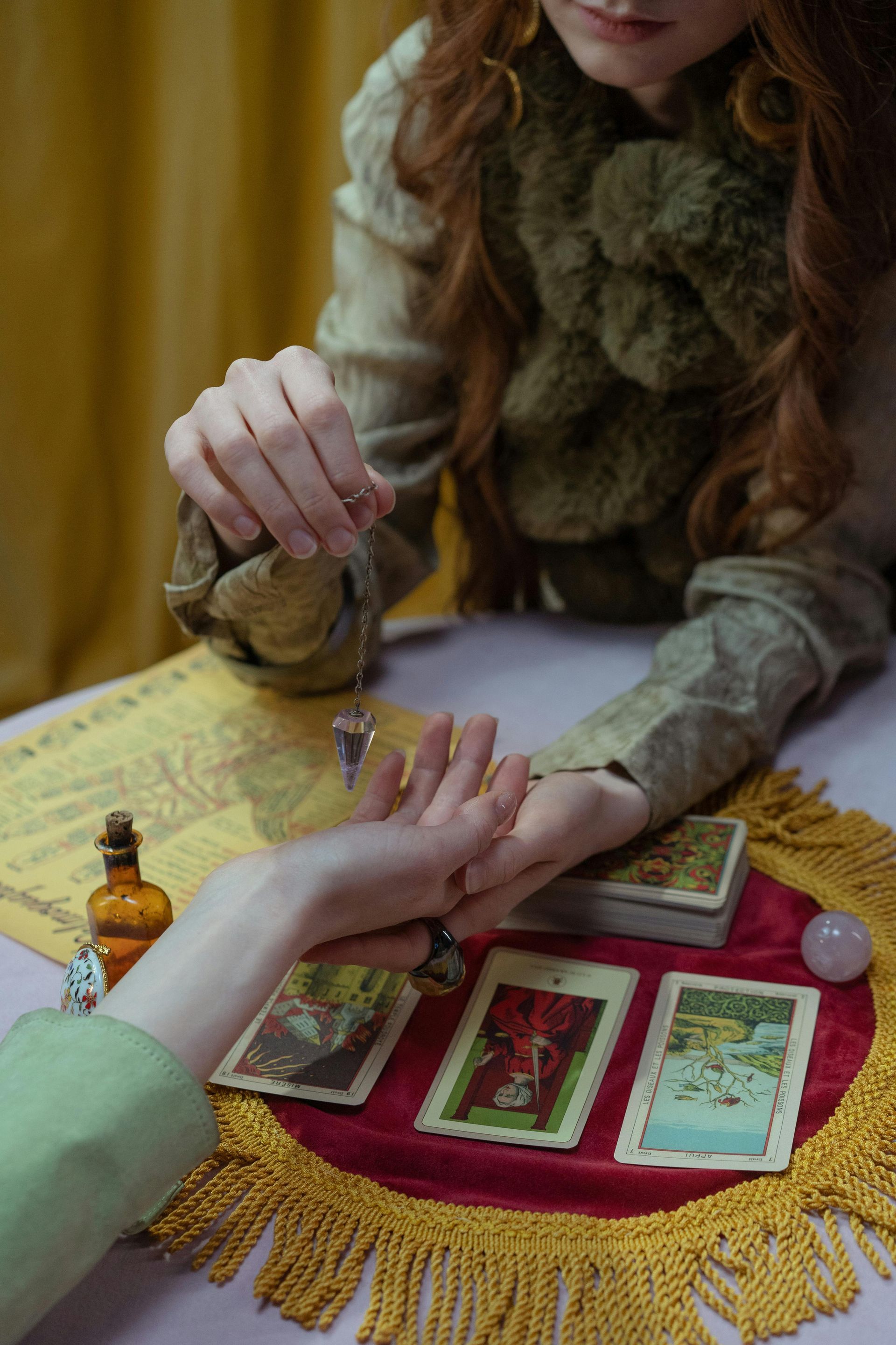 A fortune teller using a pendulum over a person's palm, cards on table, ornate setting.