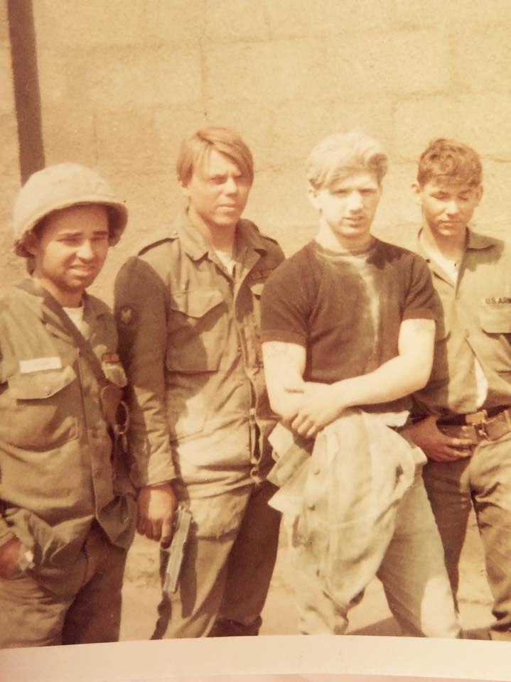 Four people, likely military personnel, stand together outdoors. Sepia-toned photo.