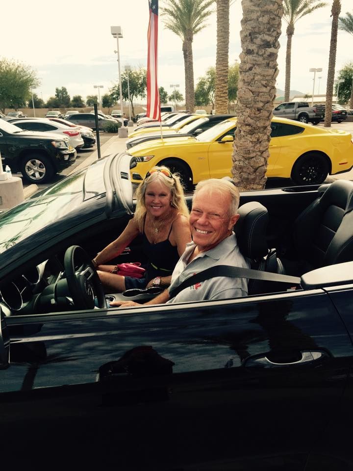 A smiling man and woman sit in a black convertible at a car dealership. Yellow cars are in the background.