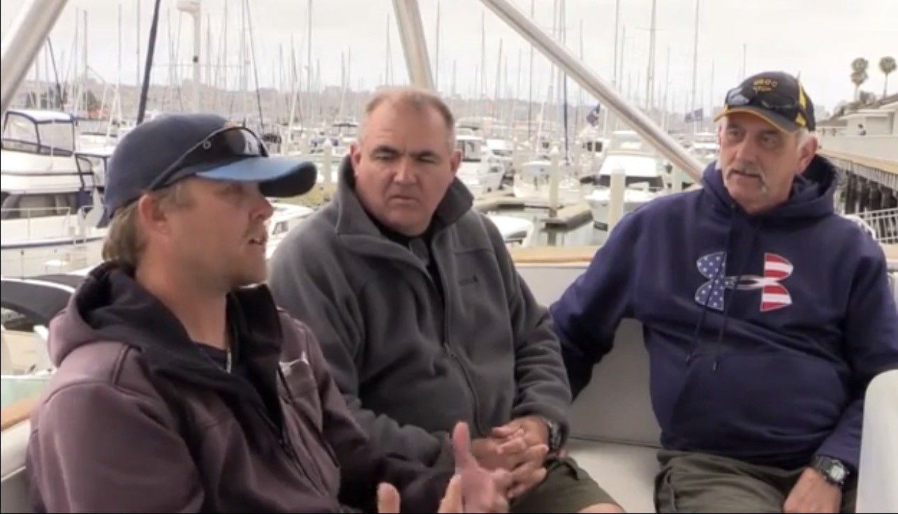 Three men seated on a boat, talking. Waterfront setting. Two men in hoodies; one wearing a cap.