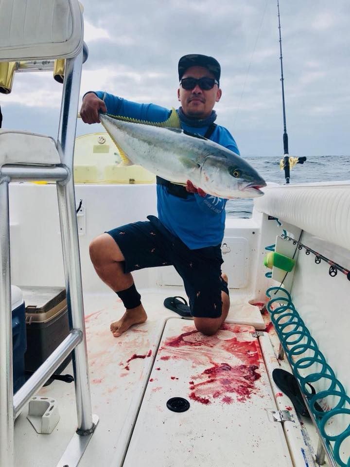 Man kneeling on boat, holding a large yellowtail fish, showing off catch. Deck shows blood. Cloudy sky.