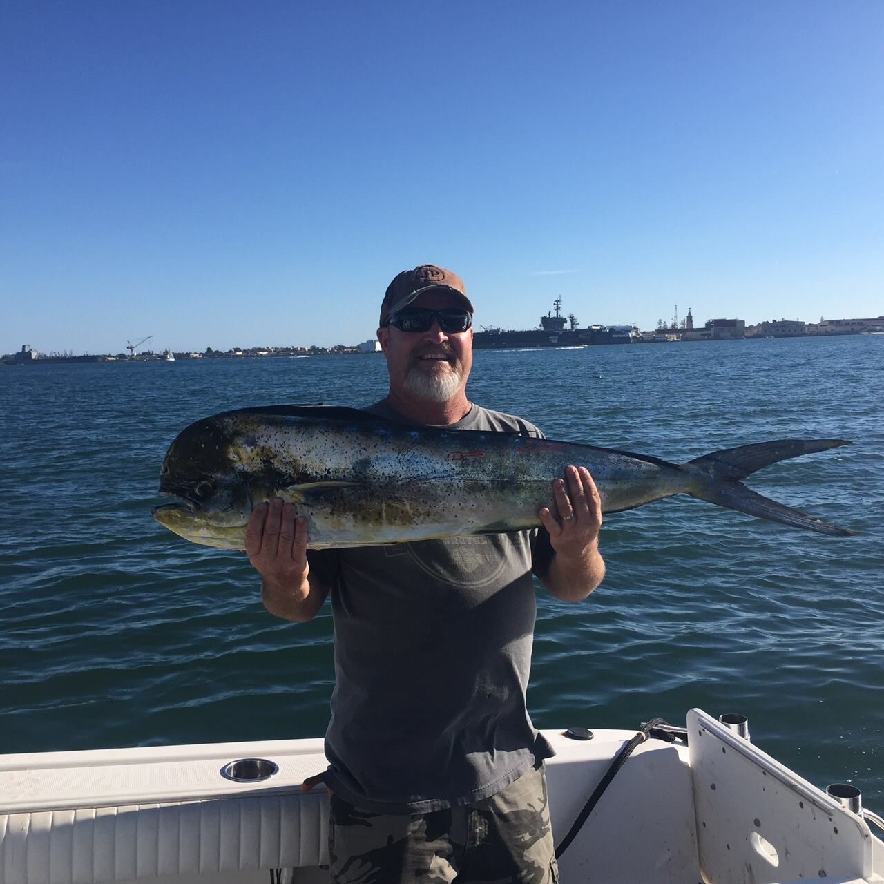 Man holding a large, silvery mahi-mahi on a boat, blue water and sky, city in the background.