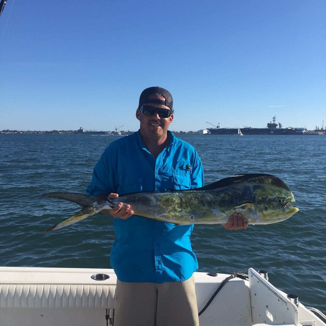 Man on a boat holds a large, colorful mahi-mahi fish; ocean and shoreline in background.