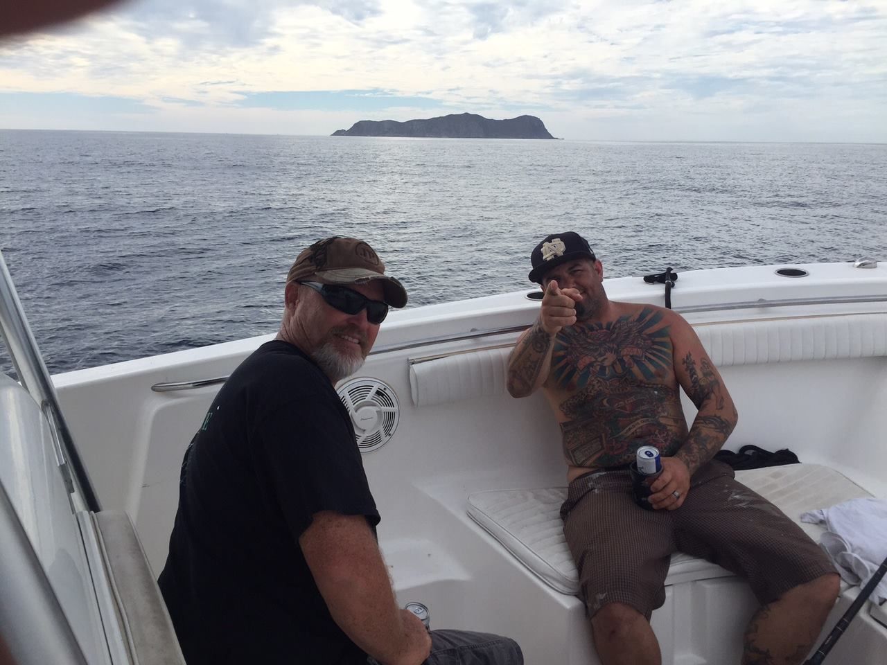 Two men on a boat, ocean view with island in the distance. One man points, the other smiles. Cloudy sky.