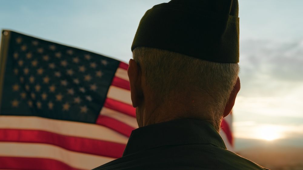 Man in uniform looks at American flag in sunset.