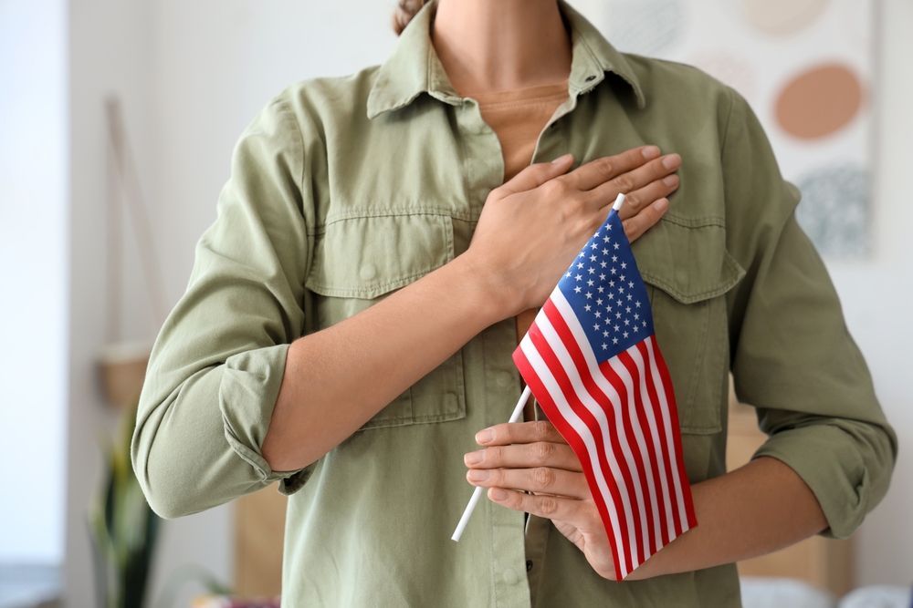 Person holding an American flag, hand on chest, indoors.
