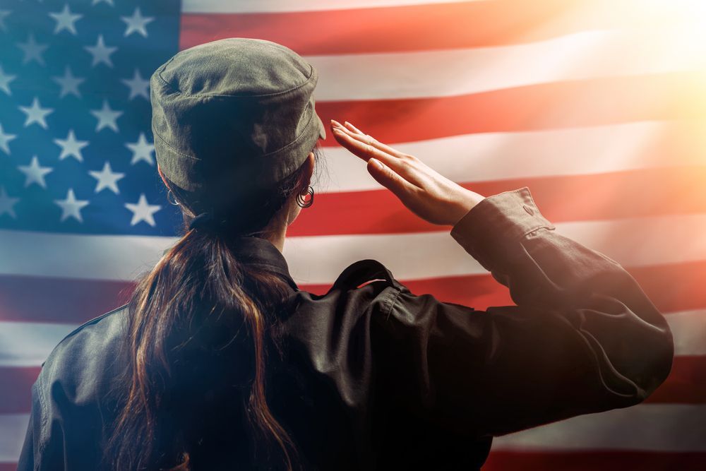 Woman in uniform saluting the American flag, backlit by sunlight.
