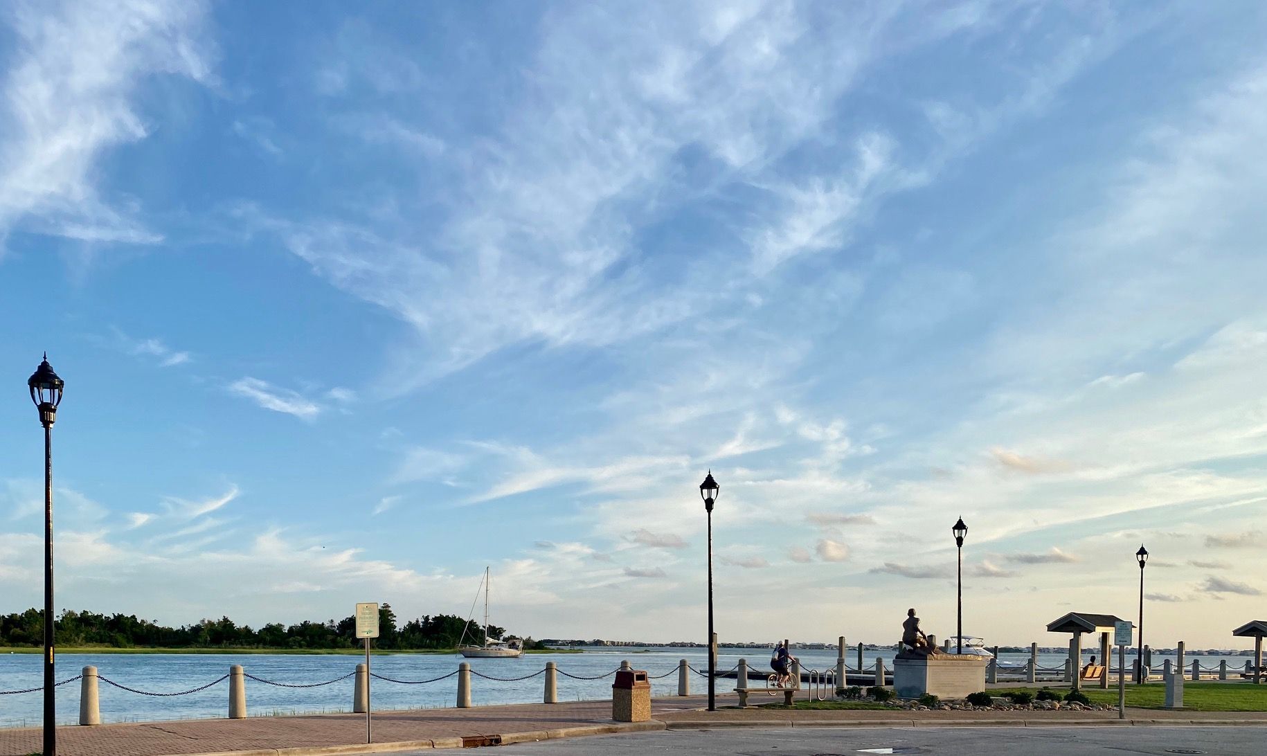 A scenic waterfront promenade with lampposts, a stone-paved walkway, and a calm river under a blue sky with wispy clouds.