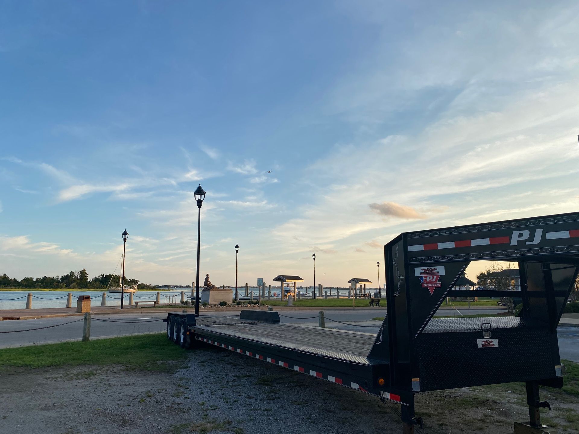 A black PJ equipment trailer parked on gravel near a waterfront park with lamp posts under a sunset sky.