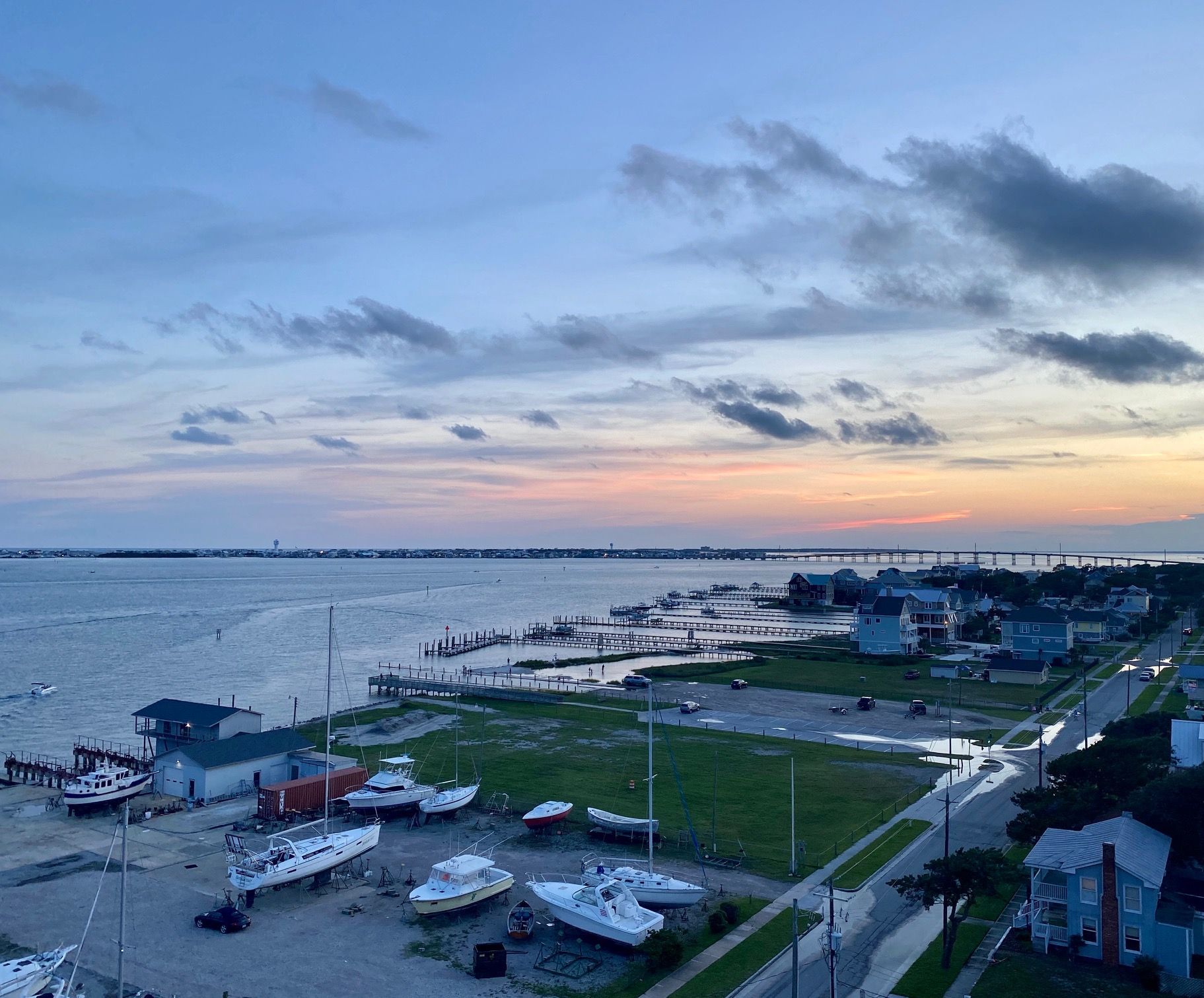 Aerial view of boats docked at a marina along a coastline during a colorful sunset, with houses and a road nearby.