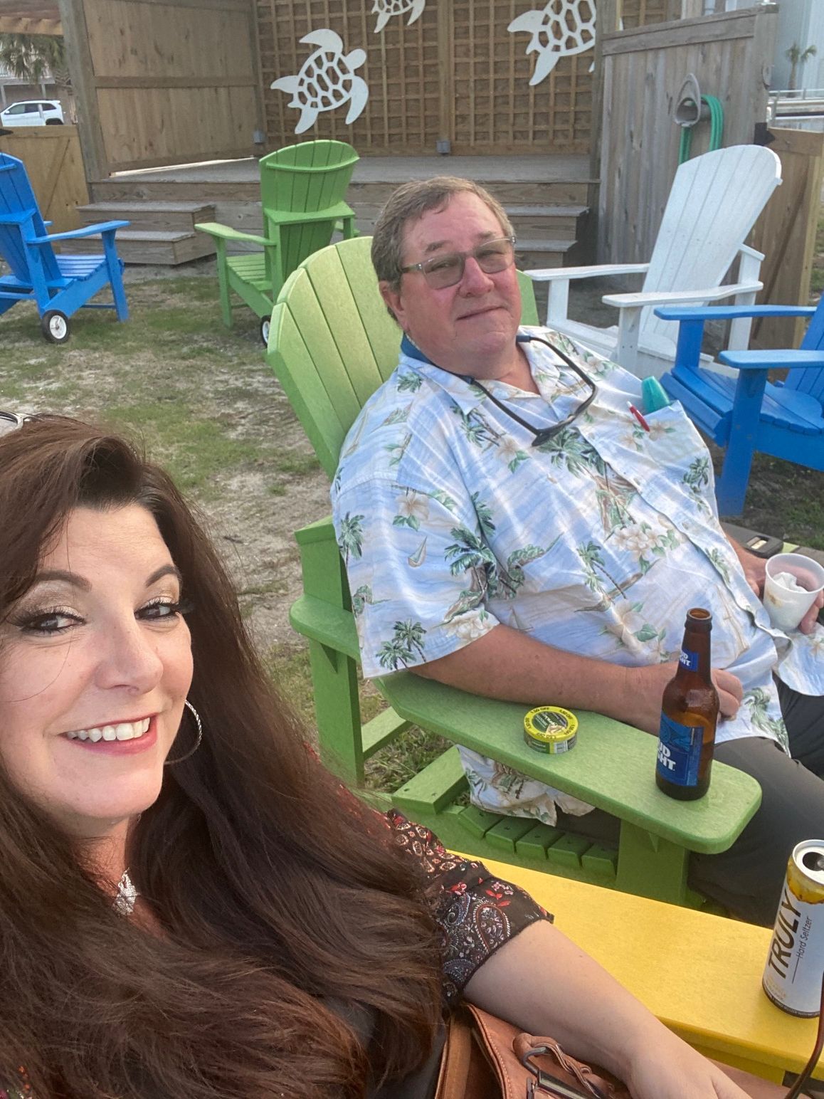 A smiling person in the foreground and a person sitting in a lime green chair with a beer, outdoors on a patio.