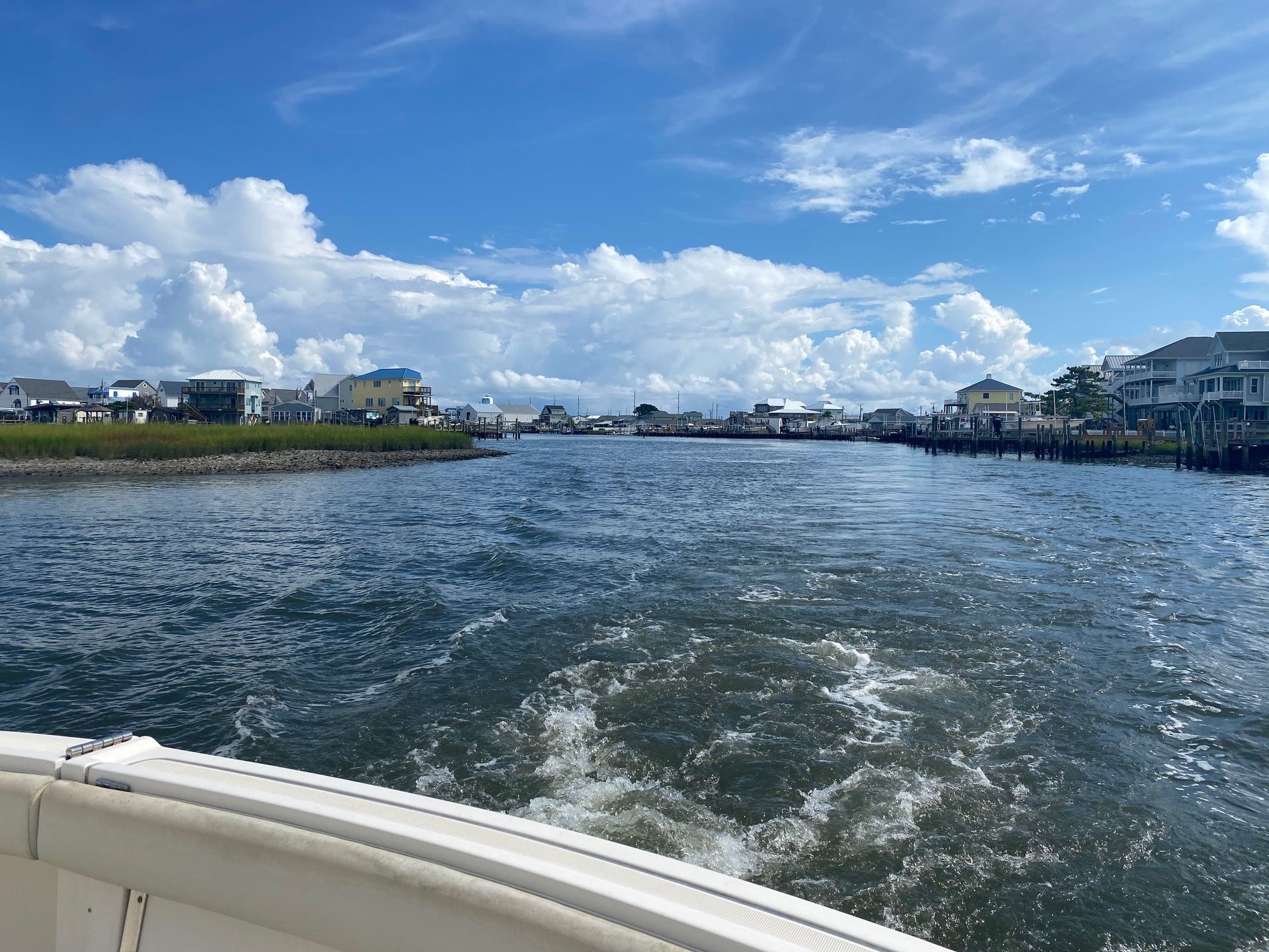 View from a boat moving through a narrow waterway toward a shoreline with houses under a bright, cloudy blue sky.