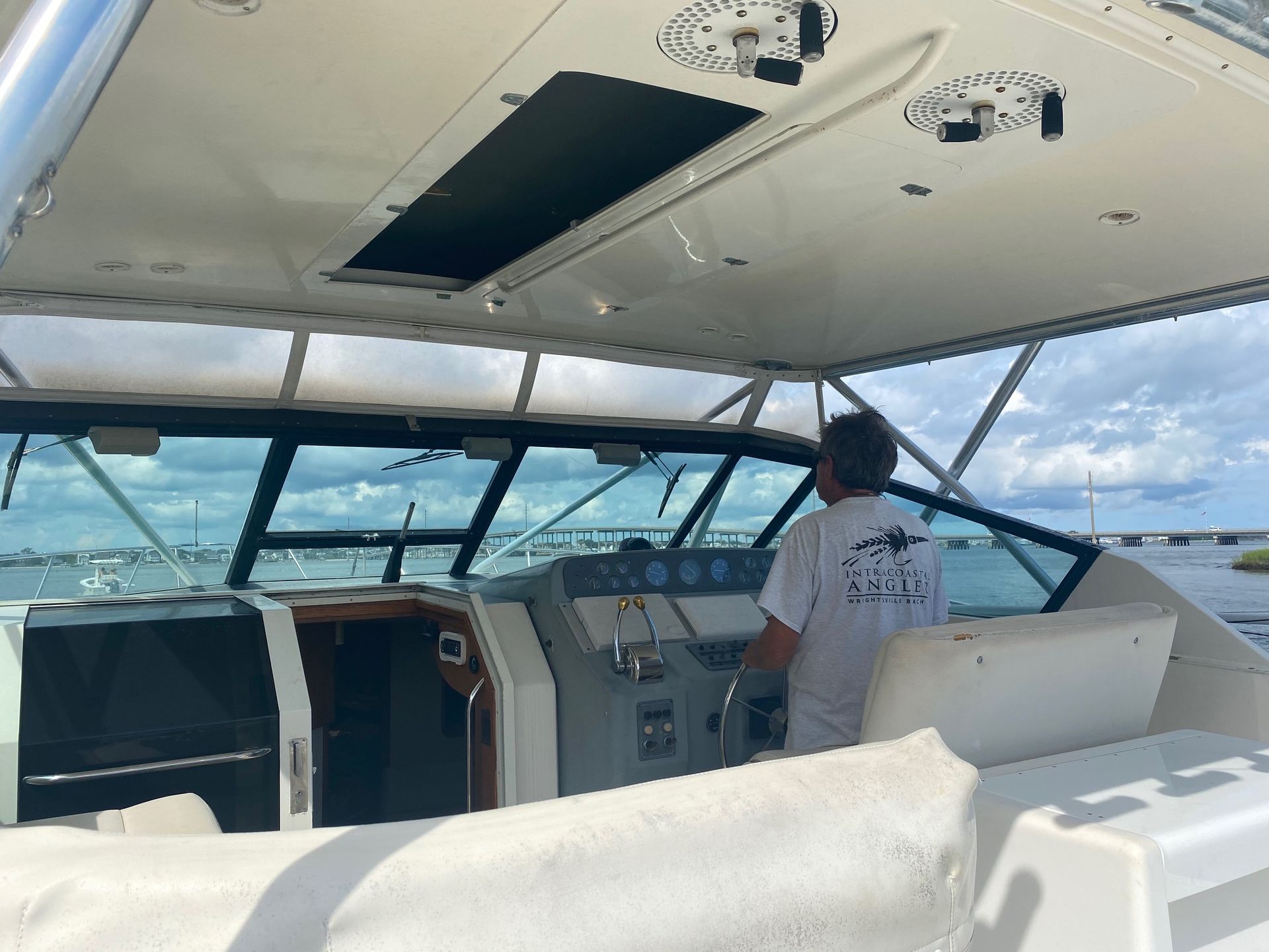 A person steers a motorboat from the helm under a hardtop cover with a view of a bridge and water in the distance.