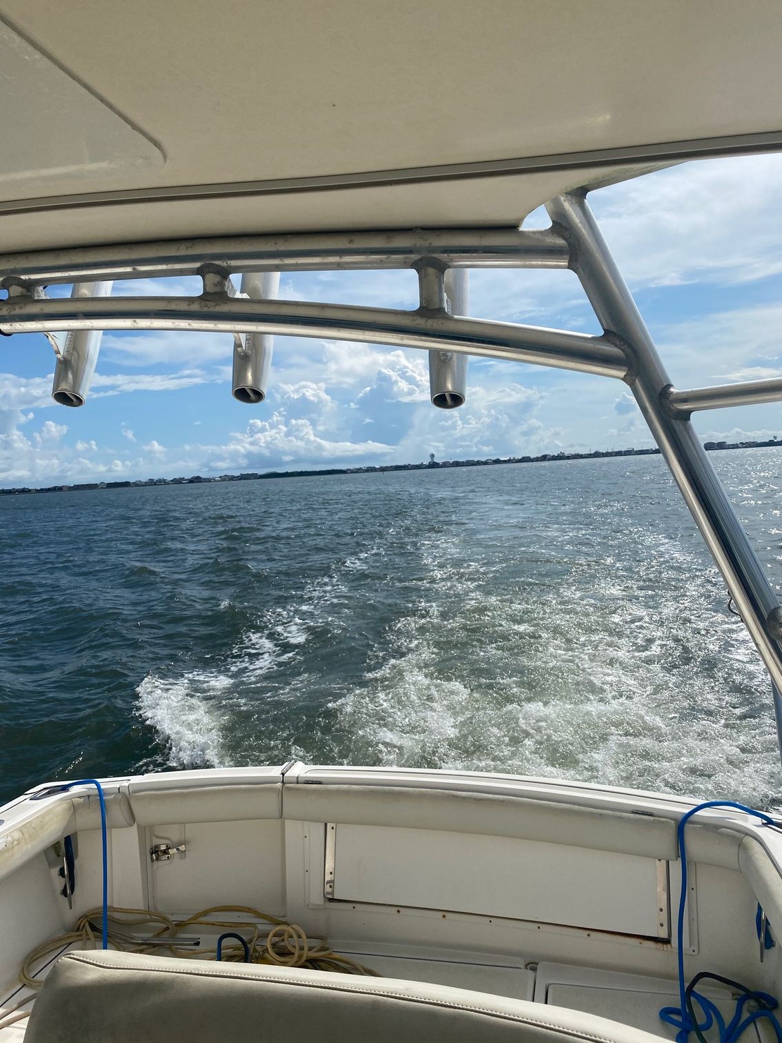 A view from the back of a moving boat, looking out over the water at a distant shoreline under a cloudy blue sky.