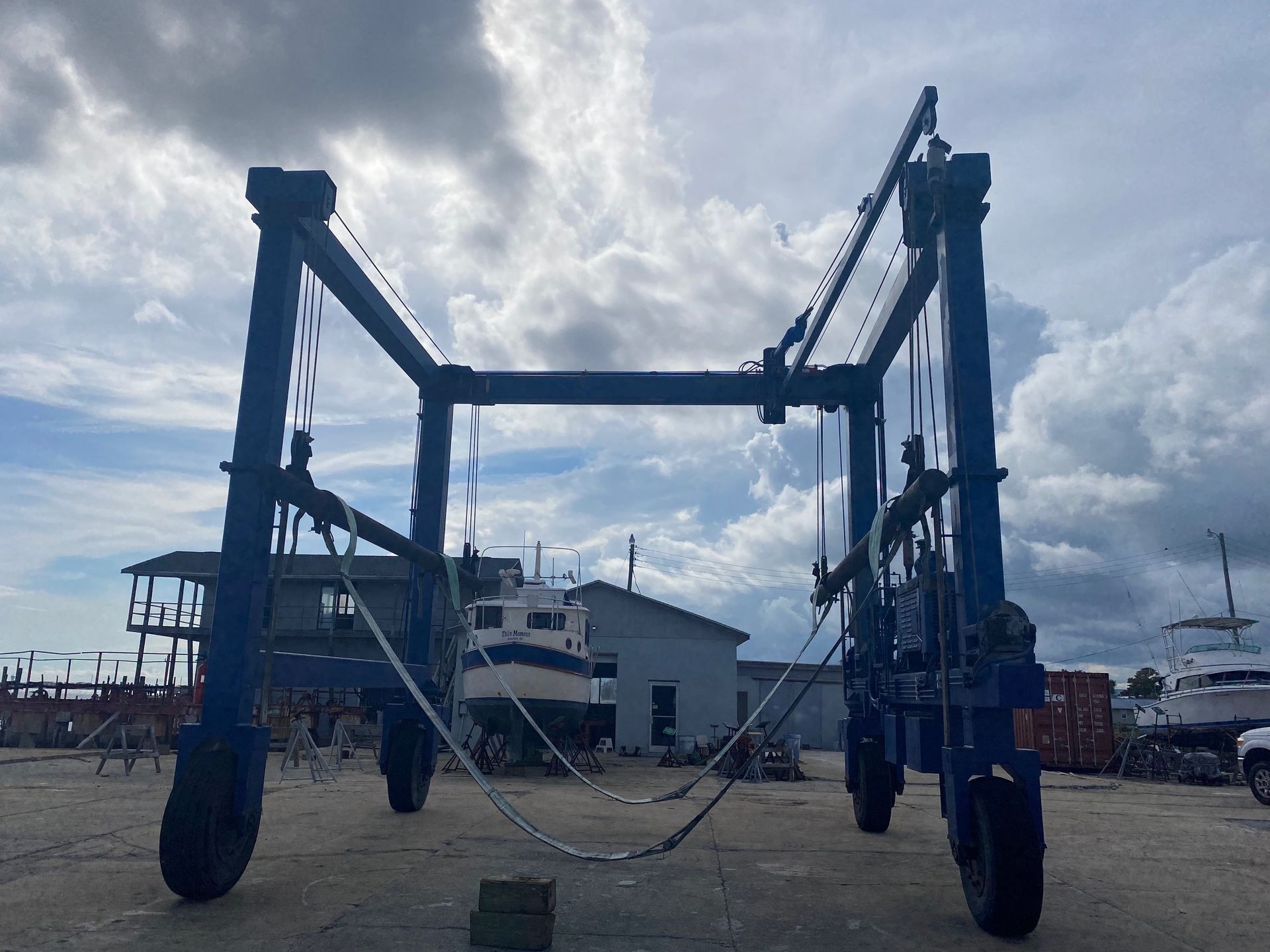 A large blue boat travel lift stands in a shipyard with a small boat visible in the background under a cloudy sky.