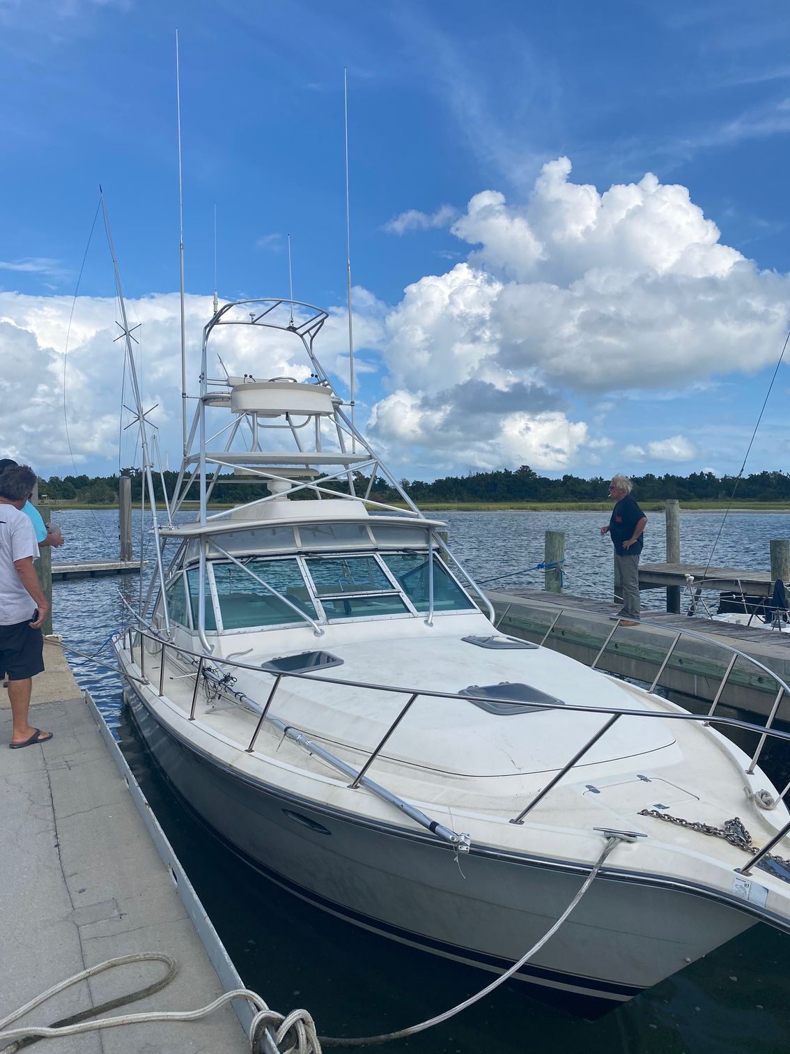 A white sport fishing boat docked at a pier under a blue sky with fluffy white clouds, with two people standing nearby.