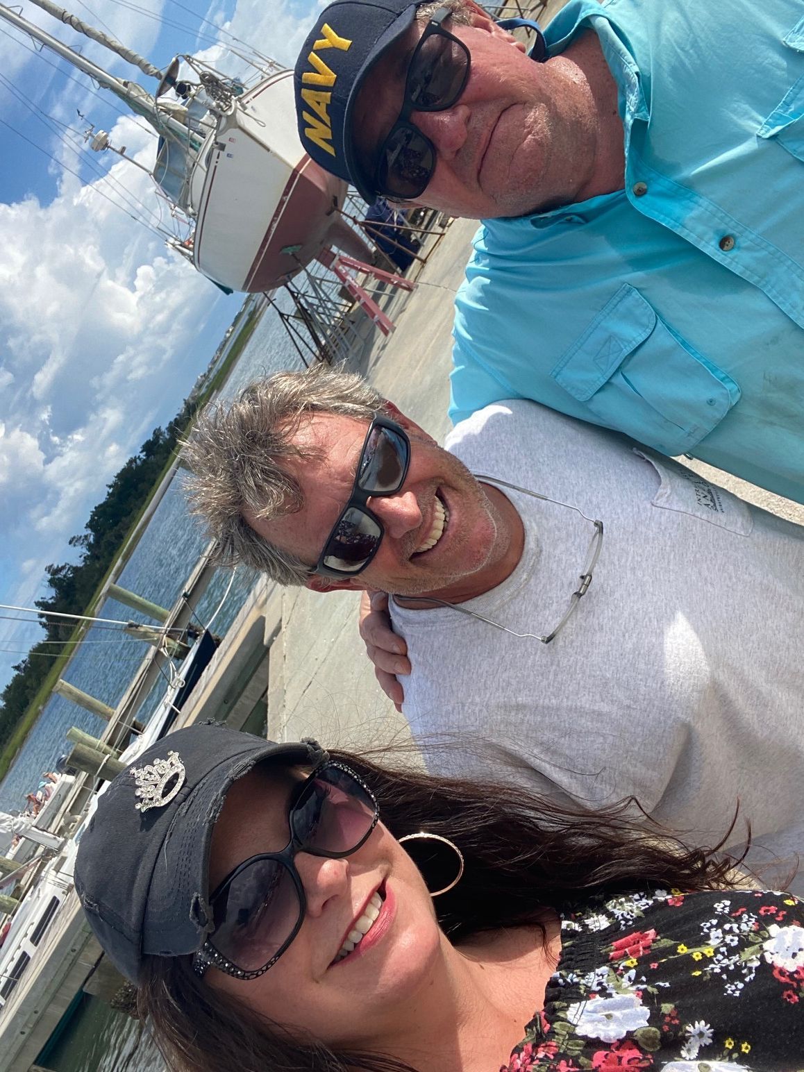 Three people wearing sunglasses pose for a selfie at a marina with boats in the background.