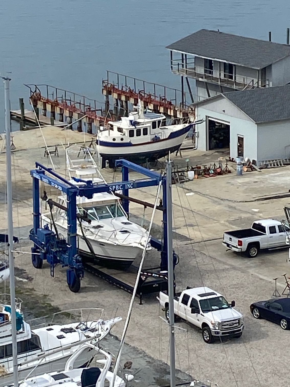 A boat lift moves a white boat across a dock, with other vessels, a building, and pickup trucks nearby in a marina setting.