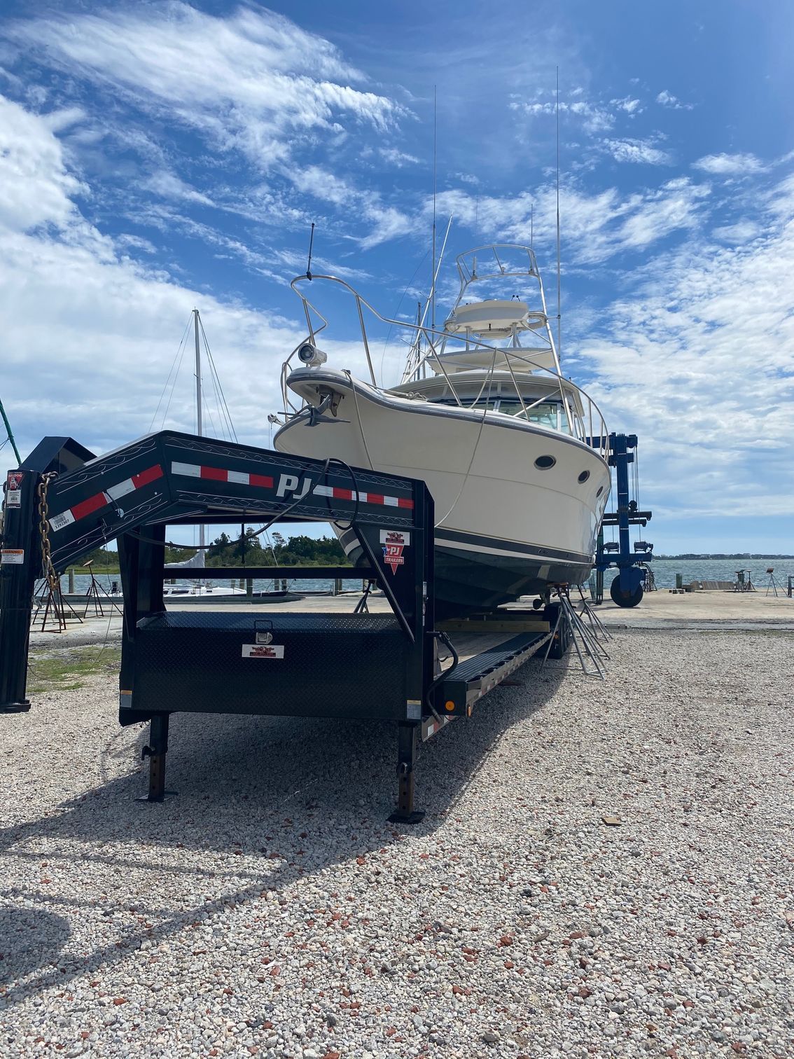 A cream-colored boat sits on a black trailer in a gravel lot under a bright, cloudy blue sky.