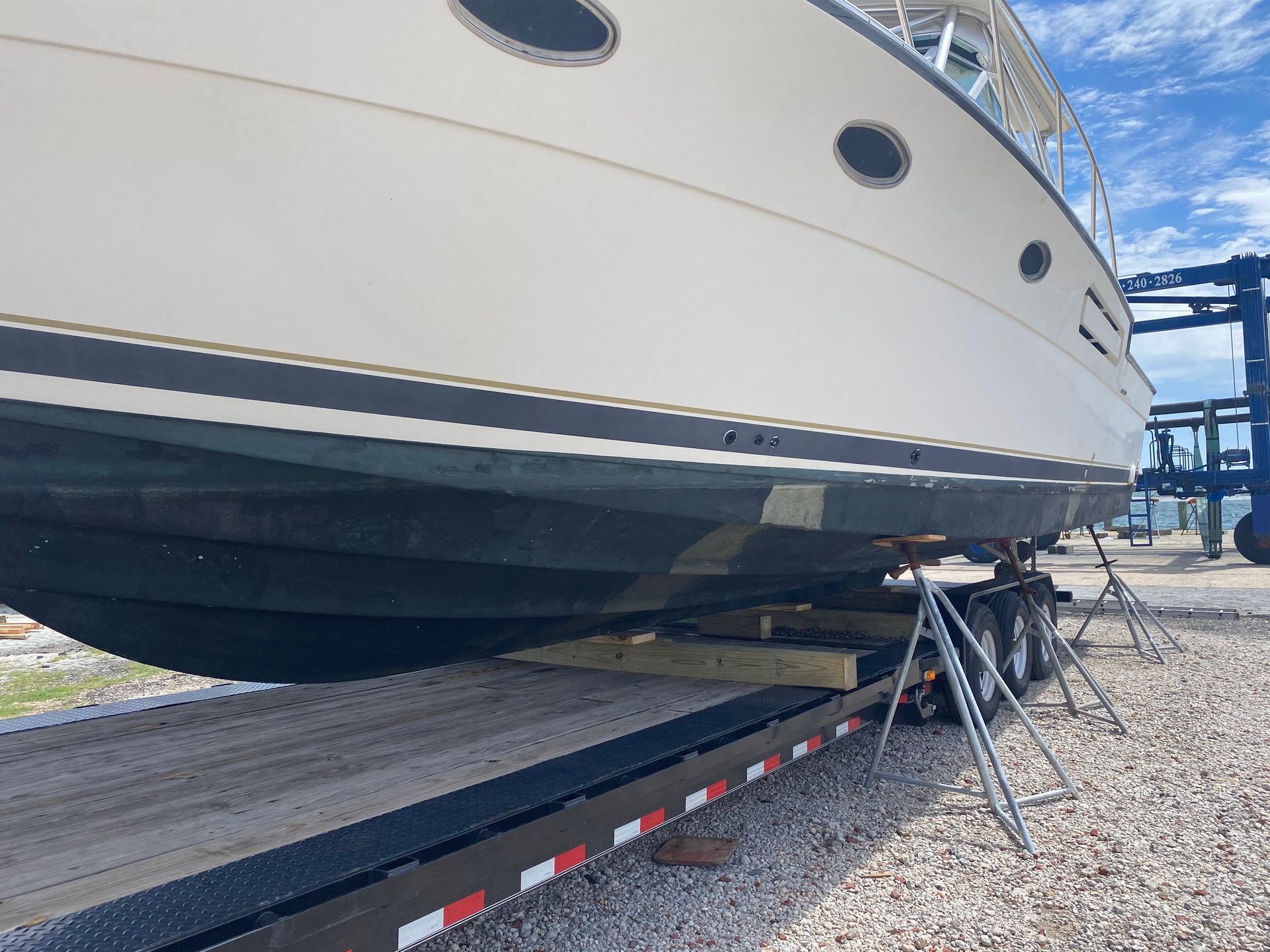 A cream-colored boat on a trailer, supported by jack stands in a gravel boat yard under a blue sky.