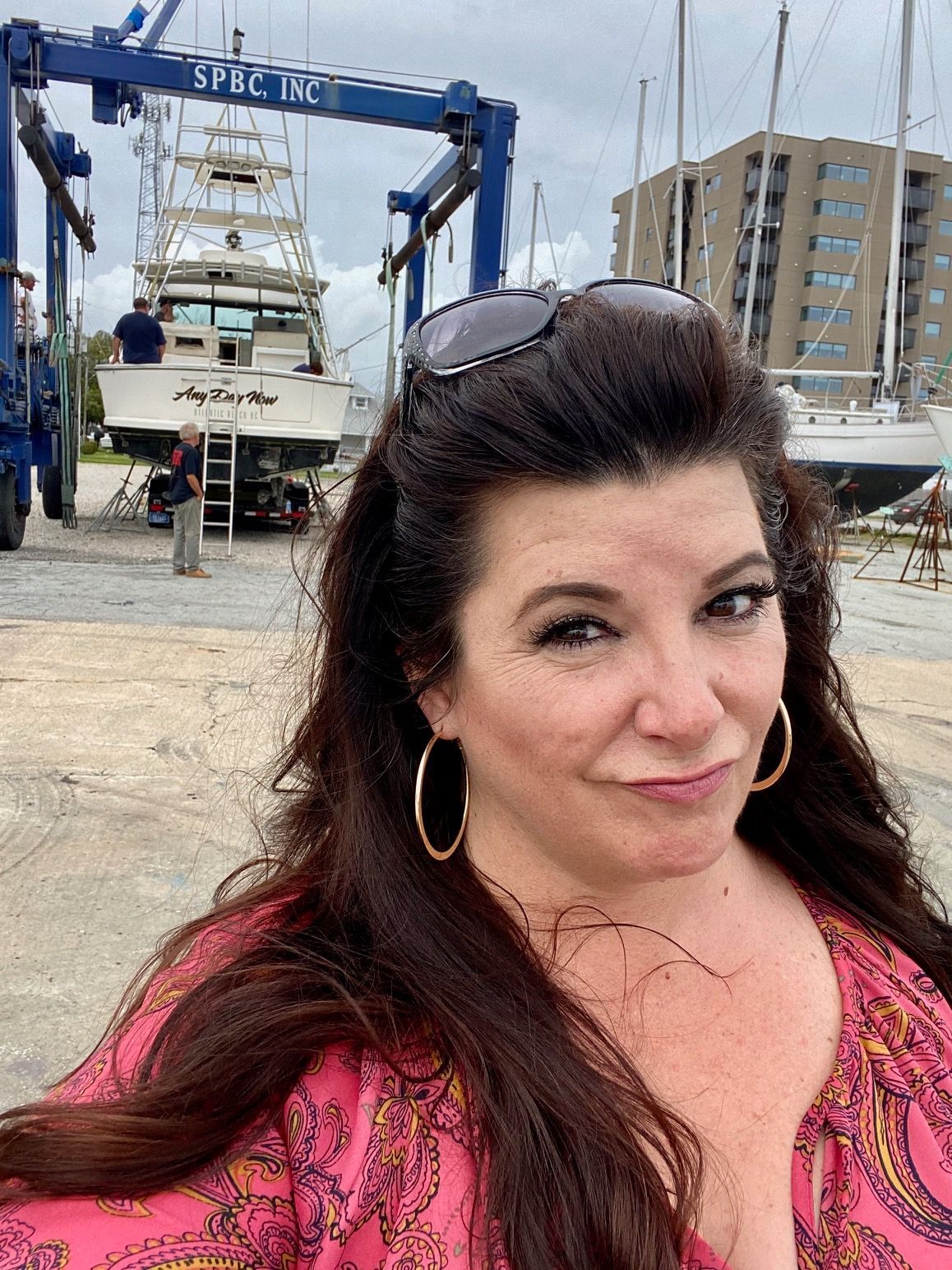 A person smiles for a selfie at a boatyard, with a large boat held by a travel lift and a building in the background.