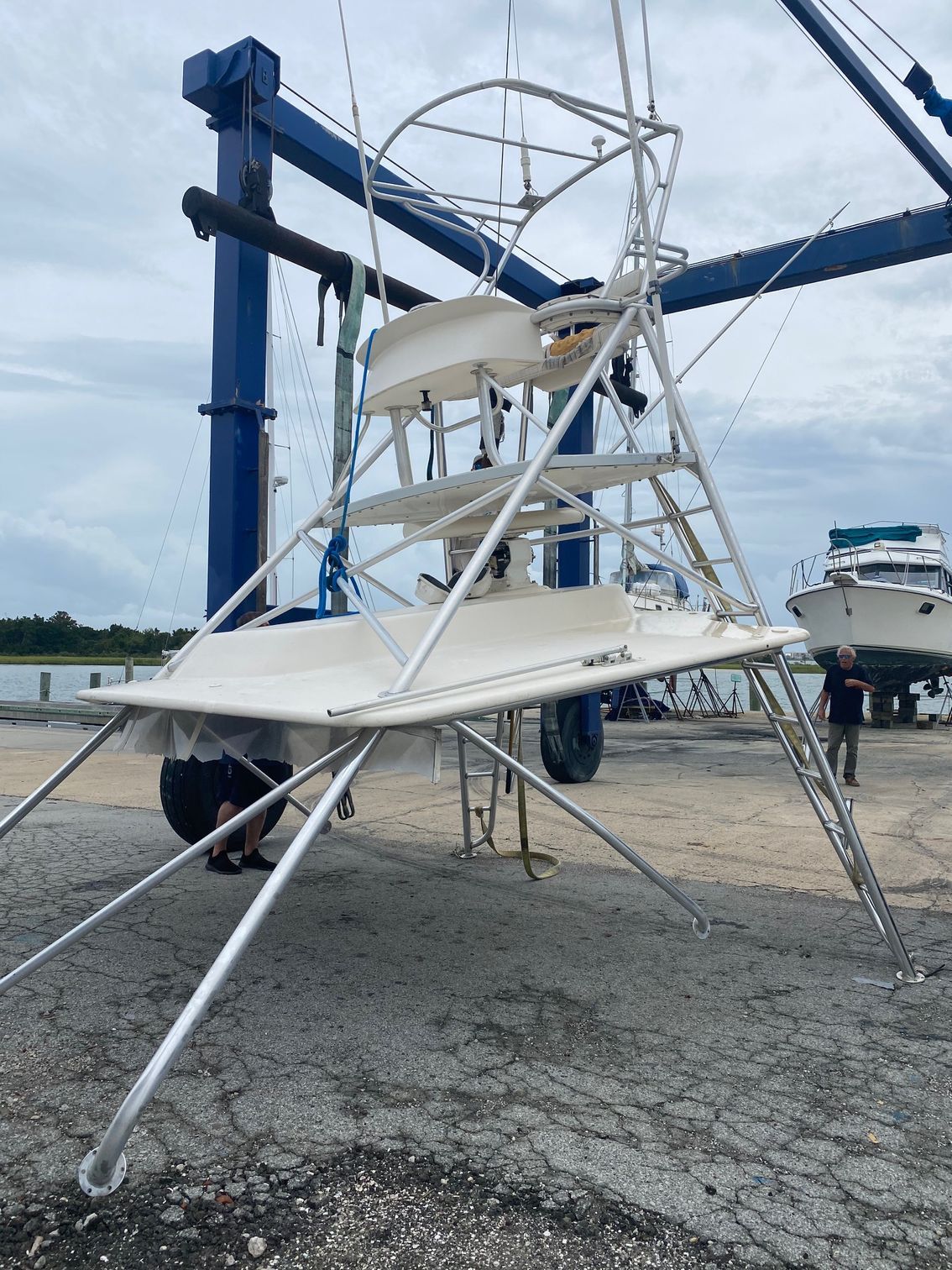 A white boat tuna tower suspended by a travel lift crane at a boat yard near the water.