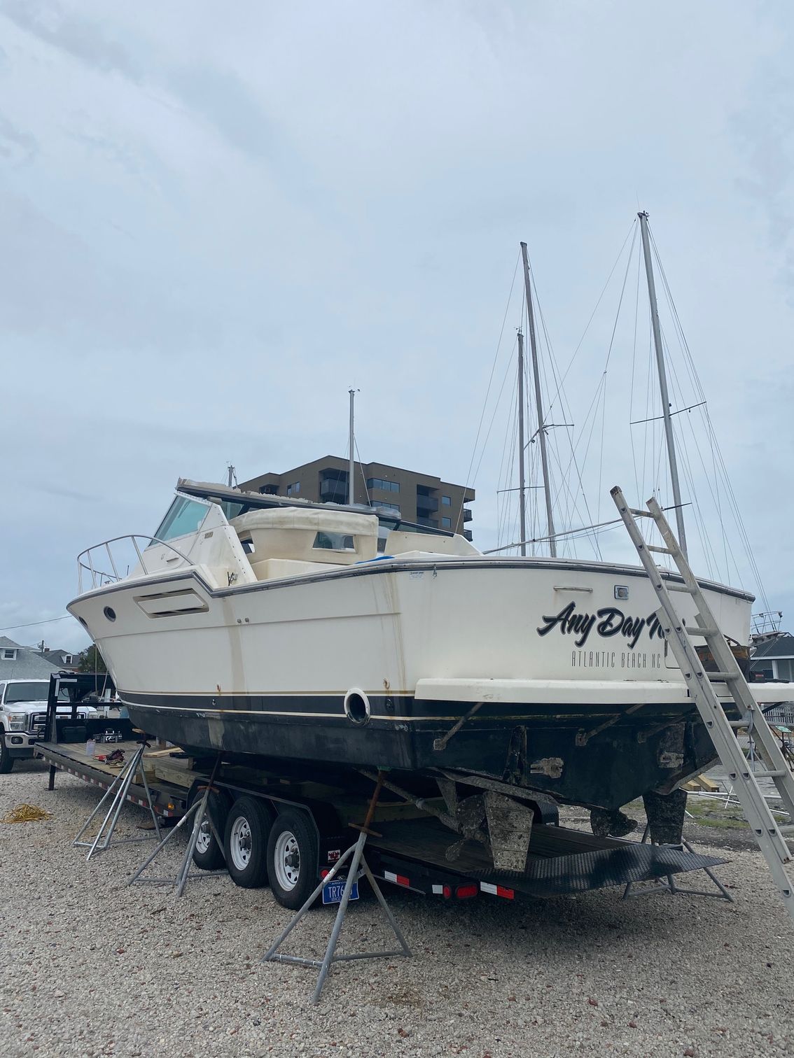 A white motorboat on a triple-axle trailer, supported by jack stands in a gravel yard, with a ladder leaning against it.