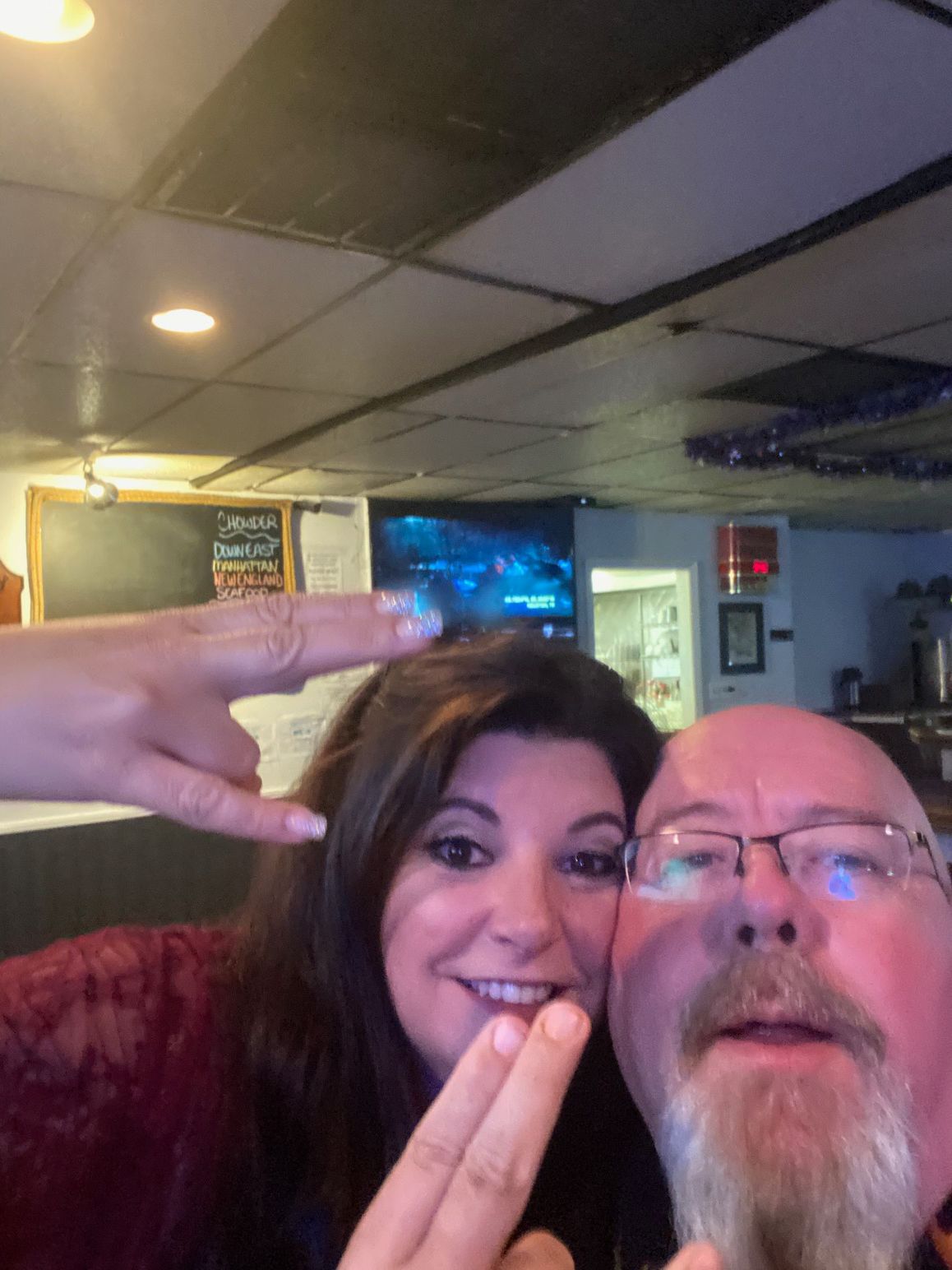 A smiling person and another person with a beard pose closely for a selfie indoors, both making hand gestures.