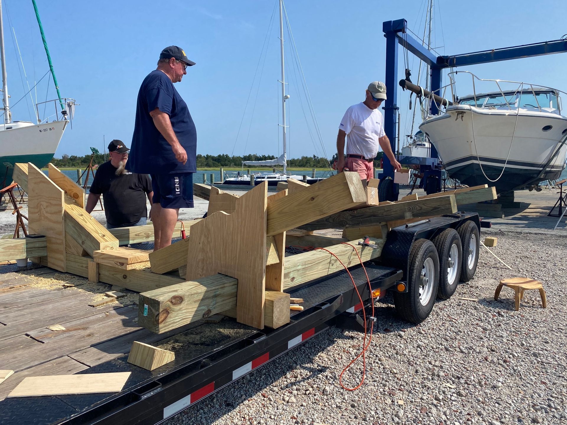 Three people assemble a custom wooden boat cradle on a trailer in a sunny boatyard near water and parked vessels.