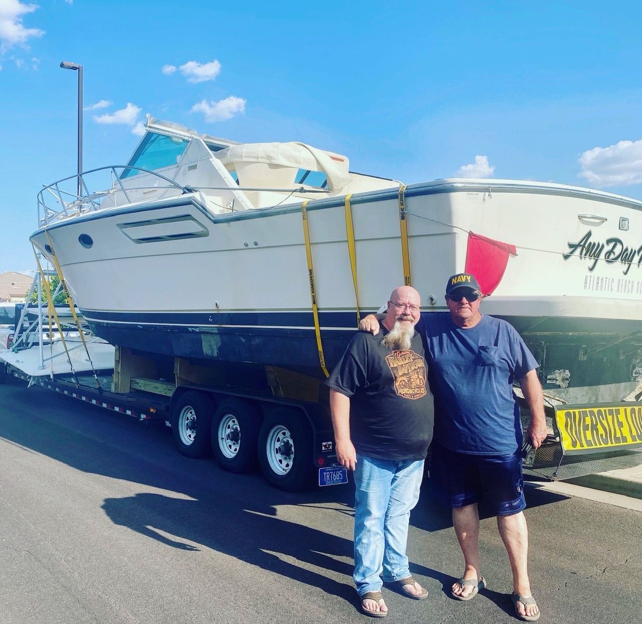 Two men stand smiling in front of a large, cream-colored cabin cruiser boat secured on a flatbed trailer.