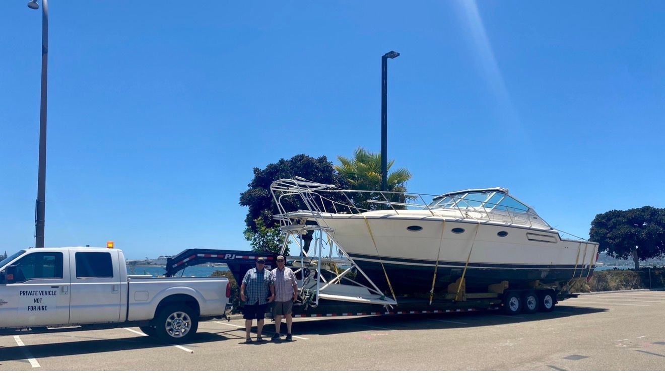 Two people stand beside a large white motorboat secured on a trailer attached to a white pickup truck in a parking lot.