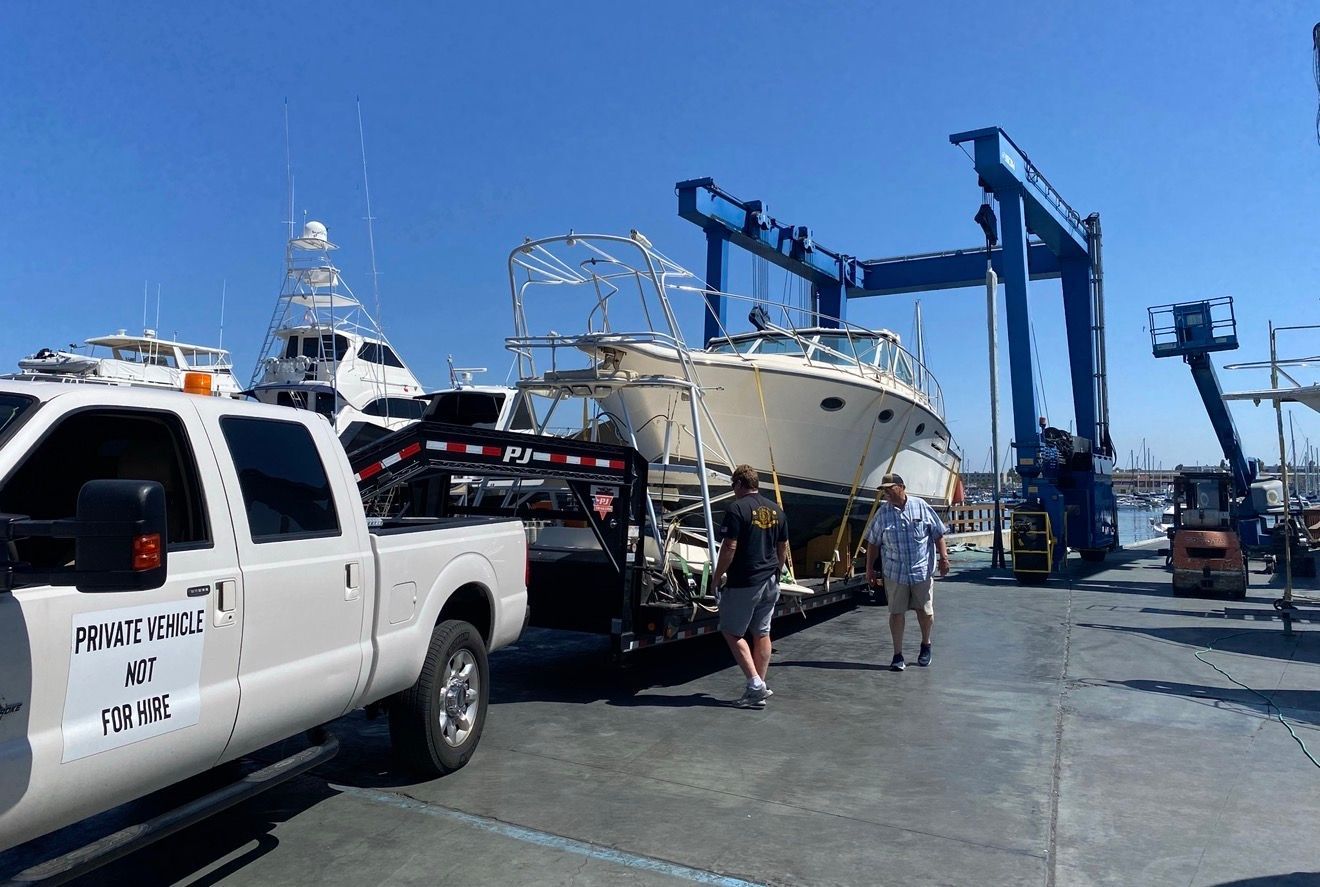 A white truck hauls a boat on a trailer past a large blue crane at a marina on a sunny day.