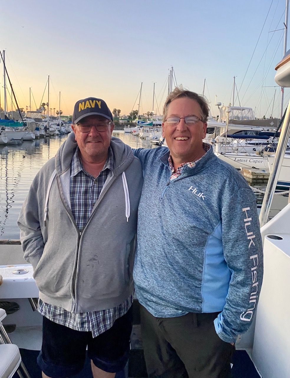 Two men stand side-by-side with arms around each other on a boat at a marina during sunset.