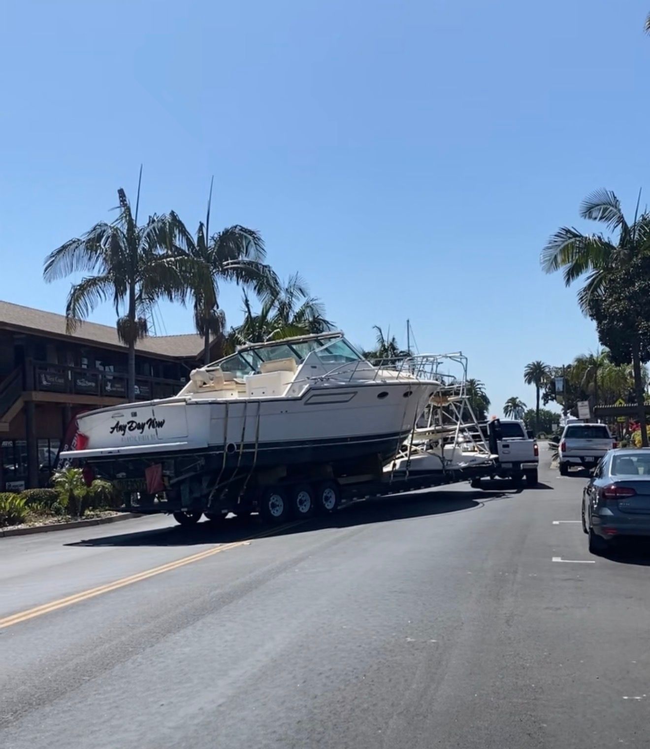 A white boat being towed on a multi-axle trailer along a sunny, palm-lined street.