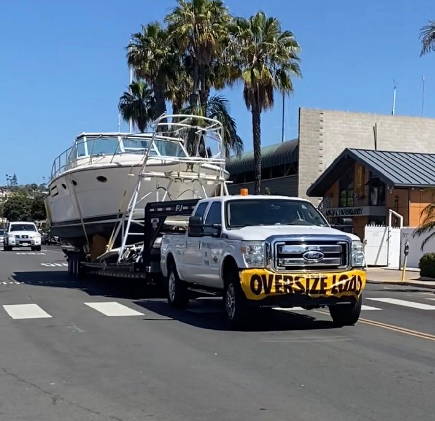 A white truck pulls a large boat on a trailer down a sunny city street, with an 